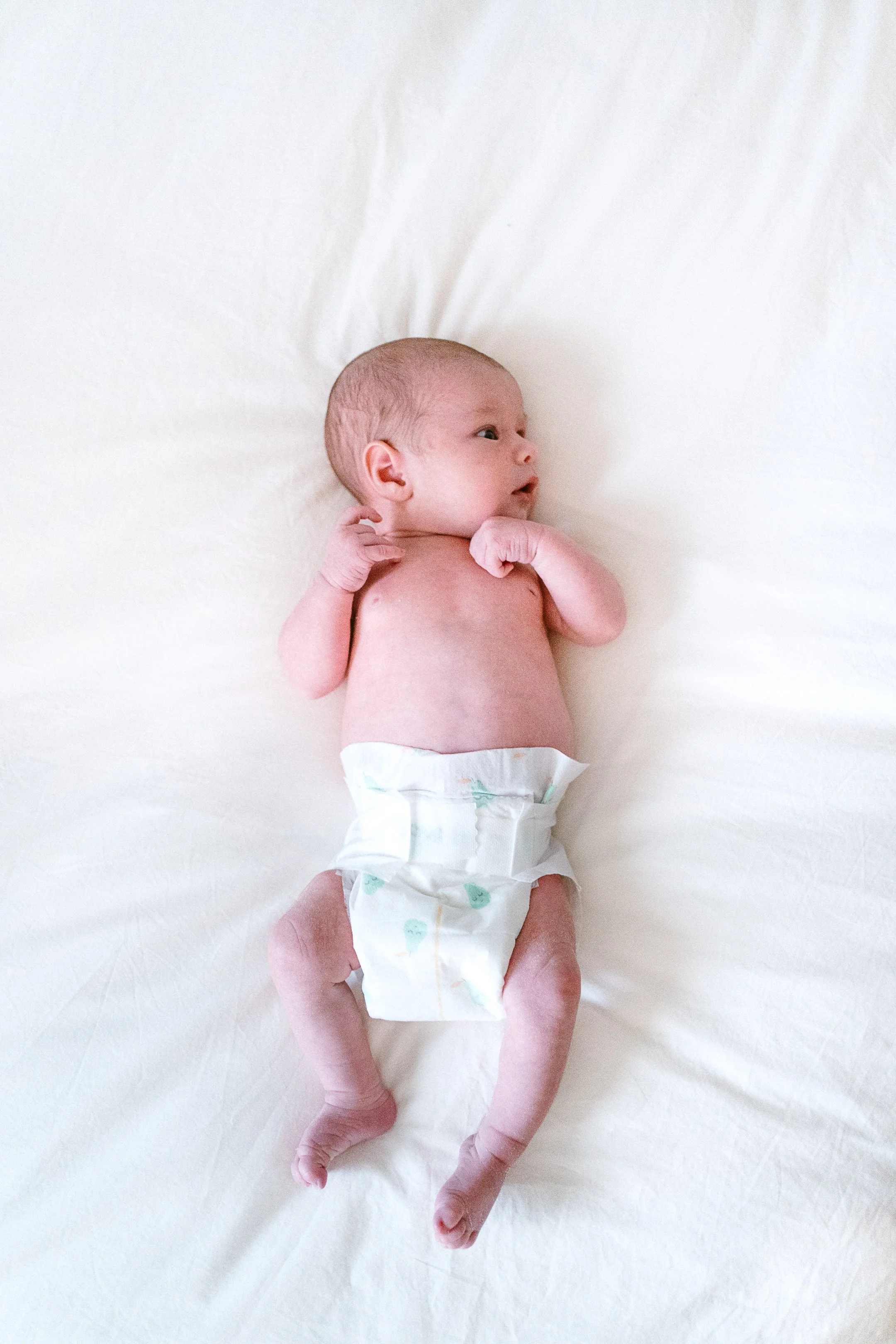A newborn baby lying on a white bed, wearing a diaper and looking to the right.