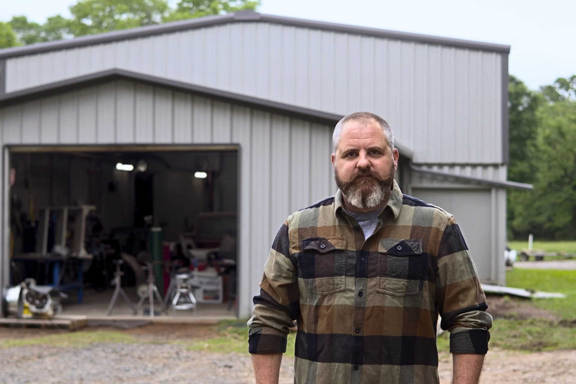 A man with a beard and mustache wearing a plaid shirt stands outside in front of a metal building with an open garage door, with trees in the background.