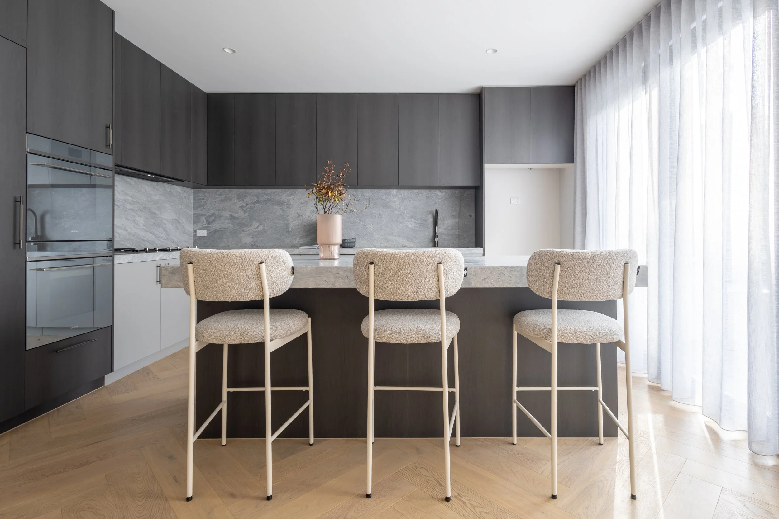 Modern kitchen with black and white cabinets, marble countertop with a vase of flowers, three beige bar stools, and a large window with sheer curtains.