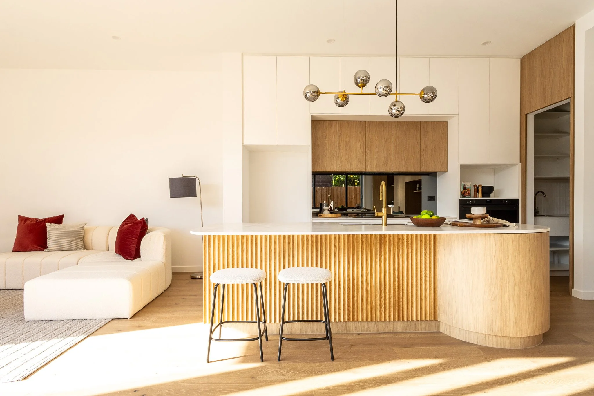 Modern open-concept living room and kitchen with white walls, wooden accents, a white sofa with red and beige pillows, a black floor lamp, and a kitchen island with a white countertop, wooden front, two white barstools, gold fixtures, and fresh limes in a wooden bowl.