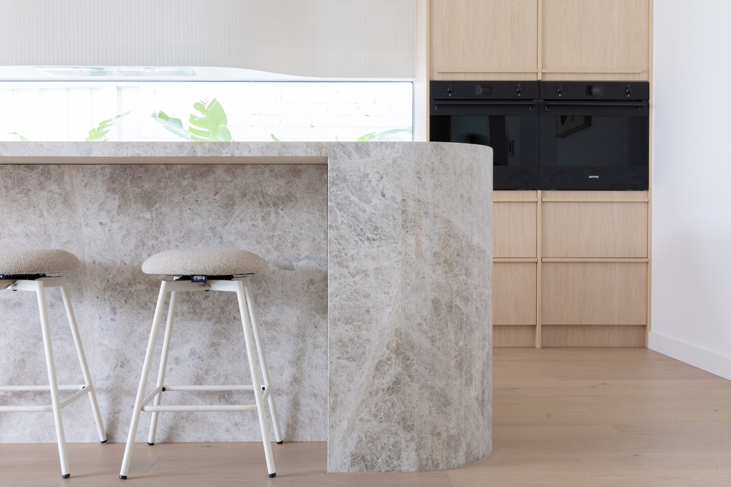 Modern kitchen with beige and gray stone island, two white bar stools with beige cushions, black built-in appliances, light wood cabinetry, and a light wood floor.