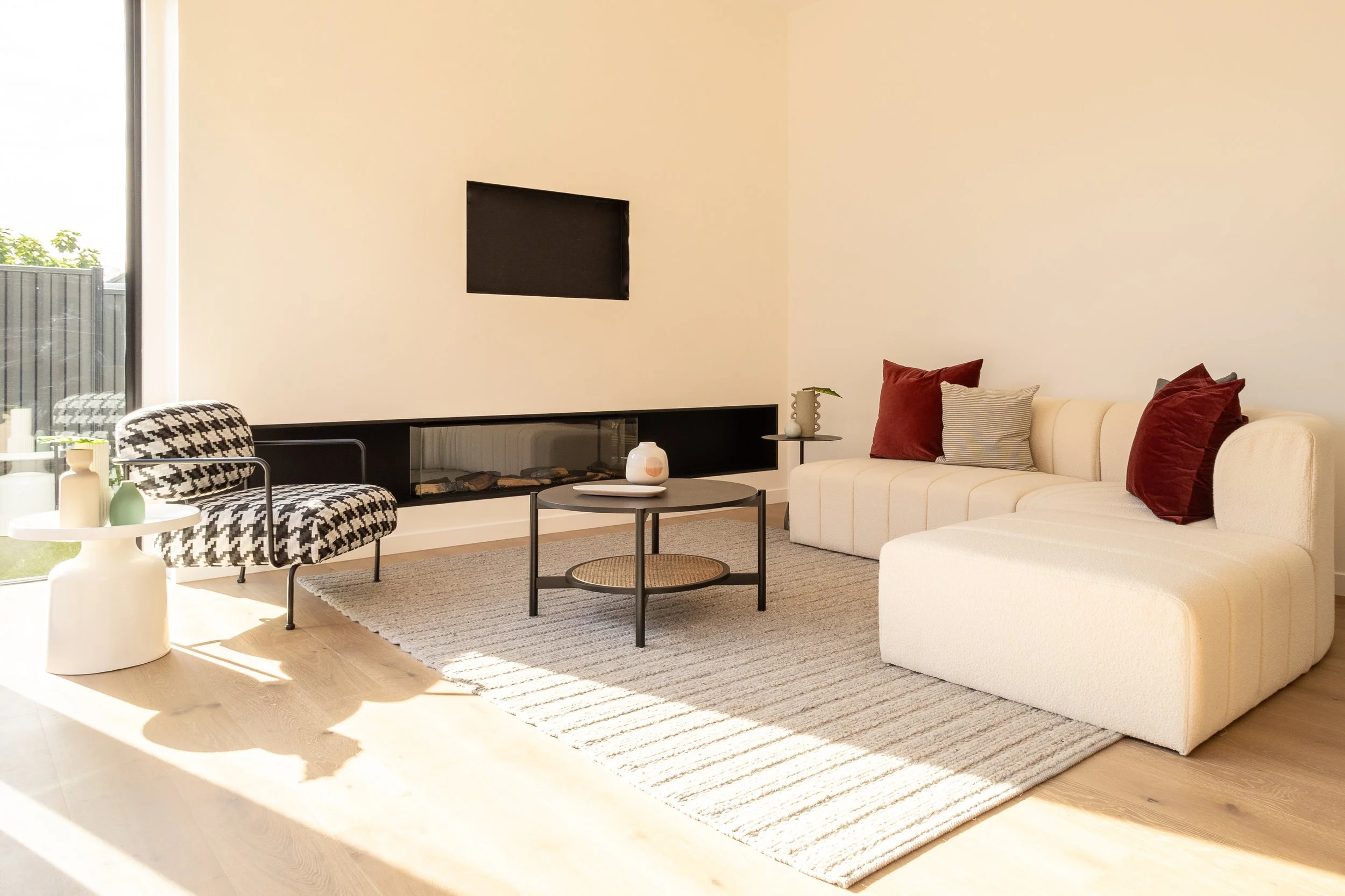 Modern living room with white sectional sofa, red and beige pillows, striped rug, black coffee table, patterned armchair, small side table with decorative items, and large window with sunlight.