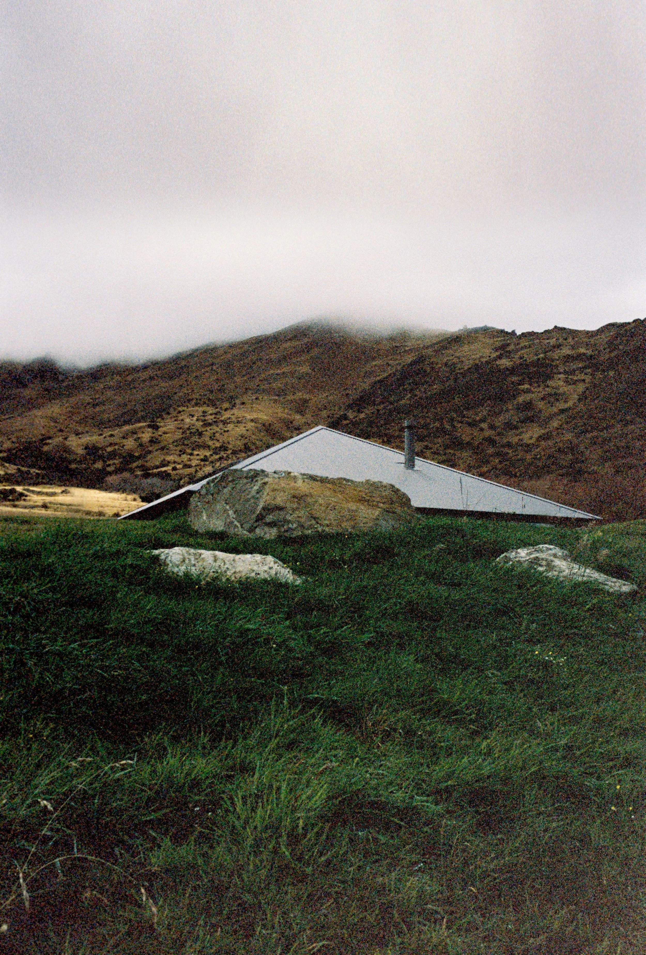 Openfield House in Queenstown, New Zealand is a house with a steep, sloped metal roof, set amidst green grass and large rocks, with a hilly, overcast landscape in the background.