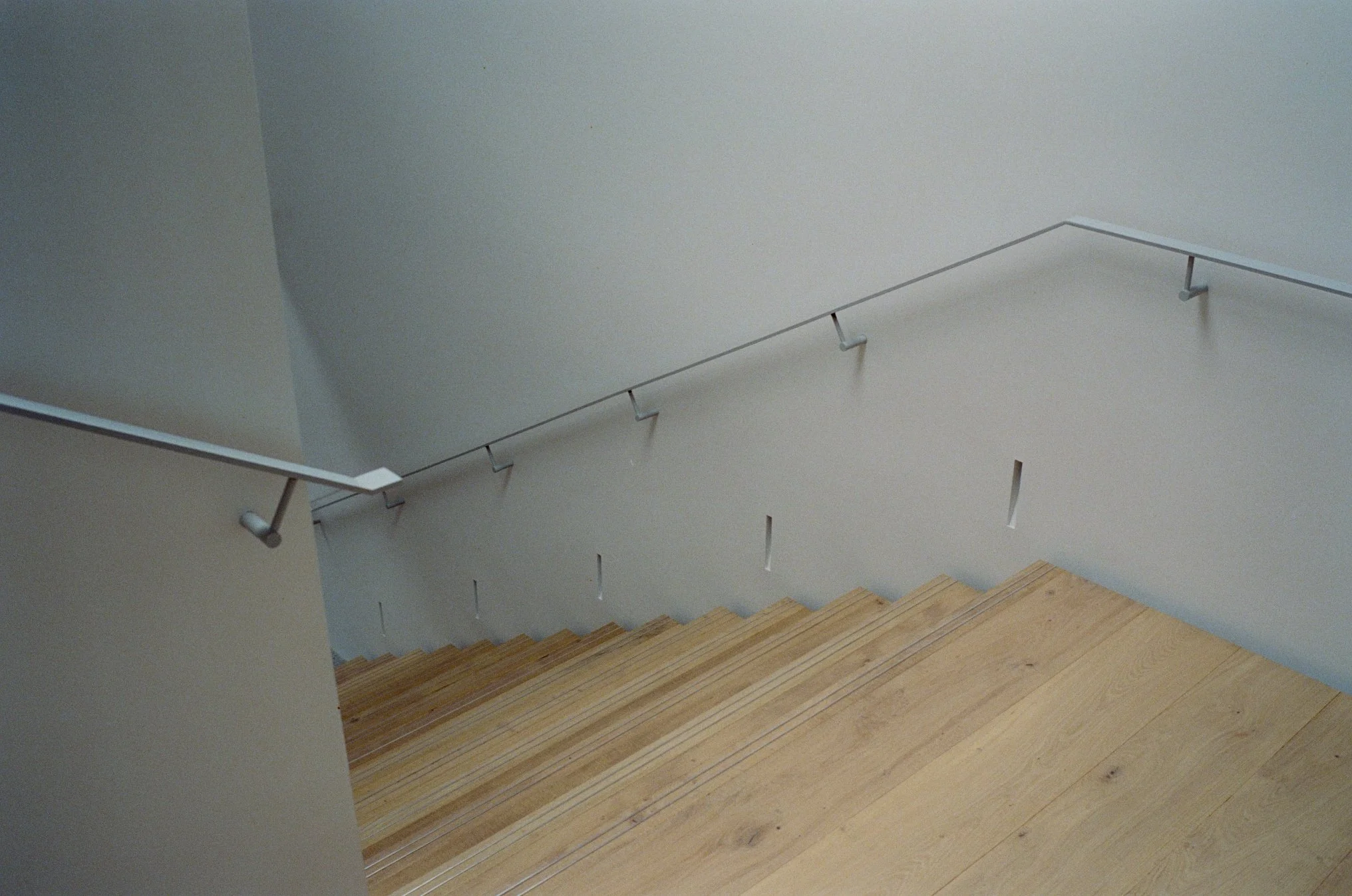 Interior view of a staircase with wooden steps and white walls, featuring a simple metal handrail mounted on the wall.