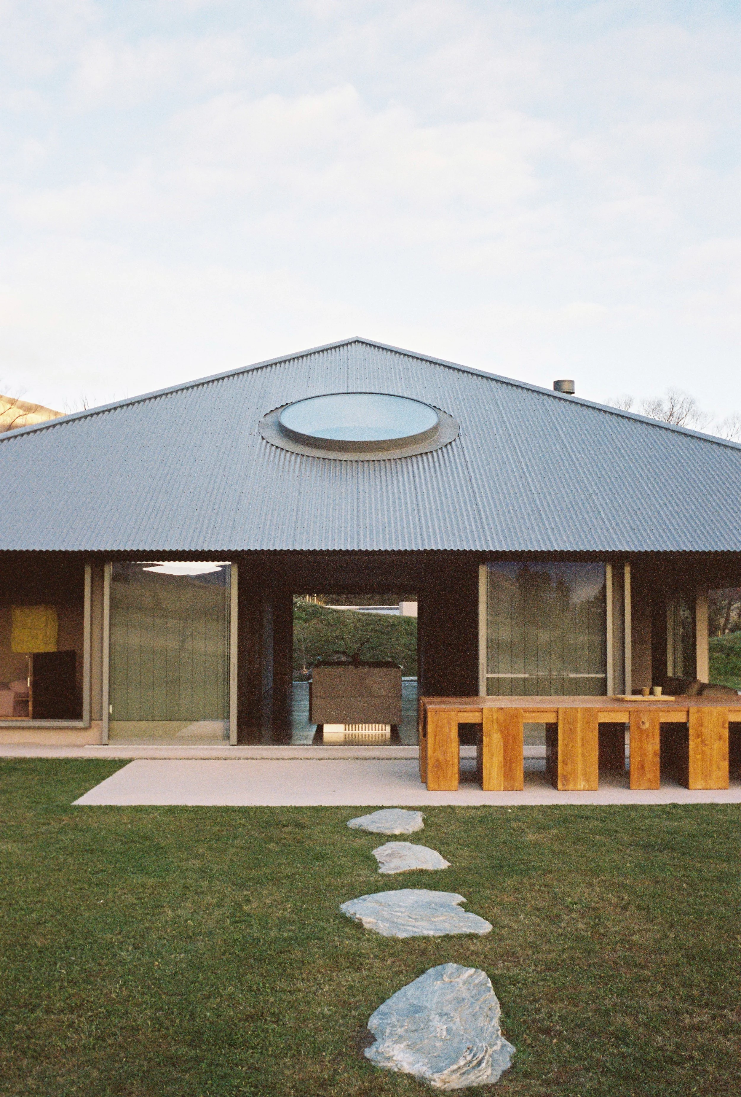 Modern house with a curved, metal roof and a round skylight, surrounded by a lawn with stepping stones leading to the entrance.