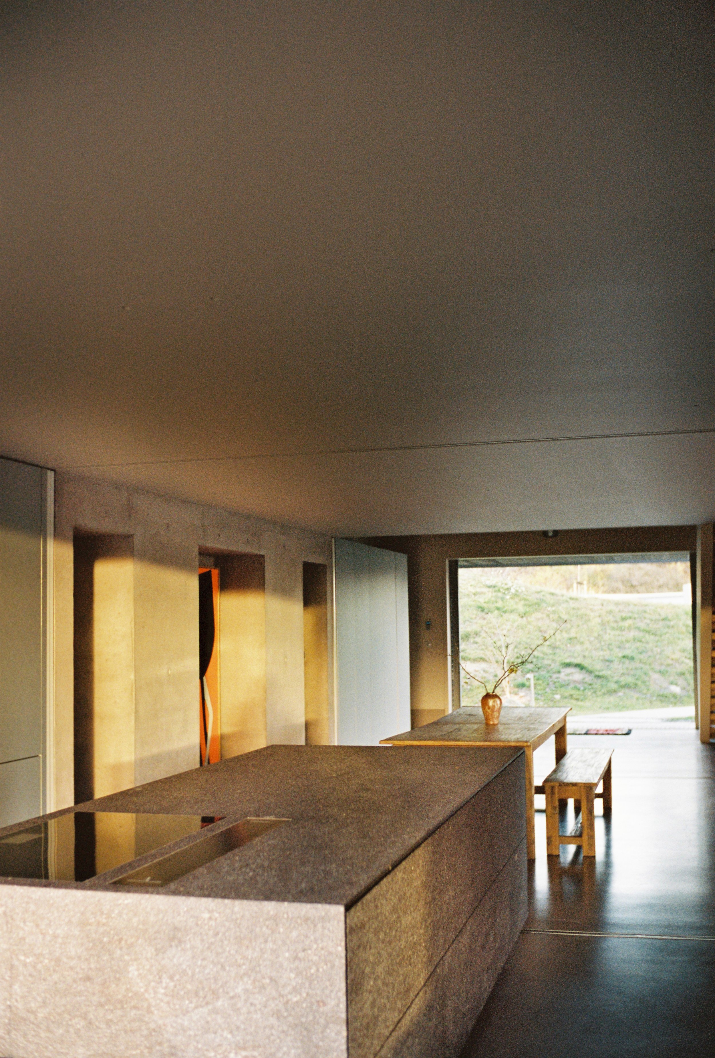 Modern kitchen with a large stone island, wooden dining table, and a vase with a branch, illuminated by warm sunlight.