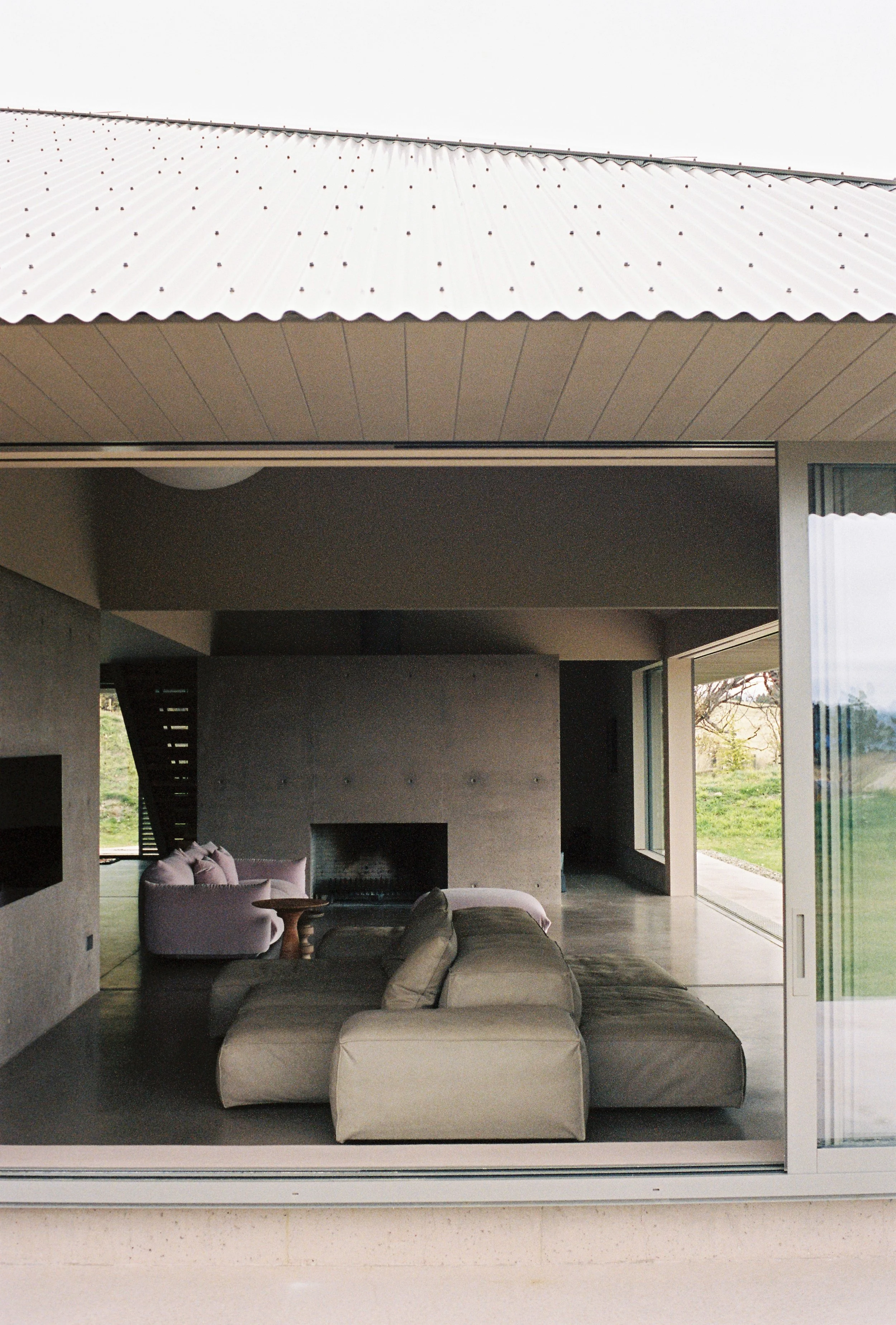 Modern living room with beige sofas, a pink armchair, a fireplace, and large windows showing a green outdoor landscape.