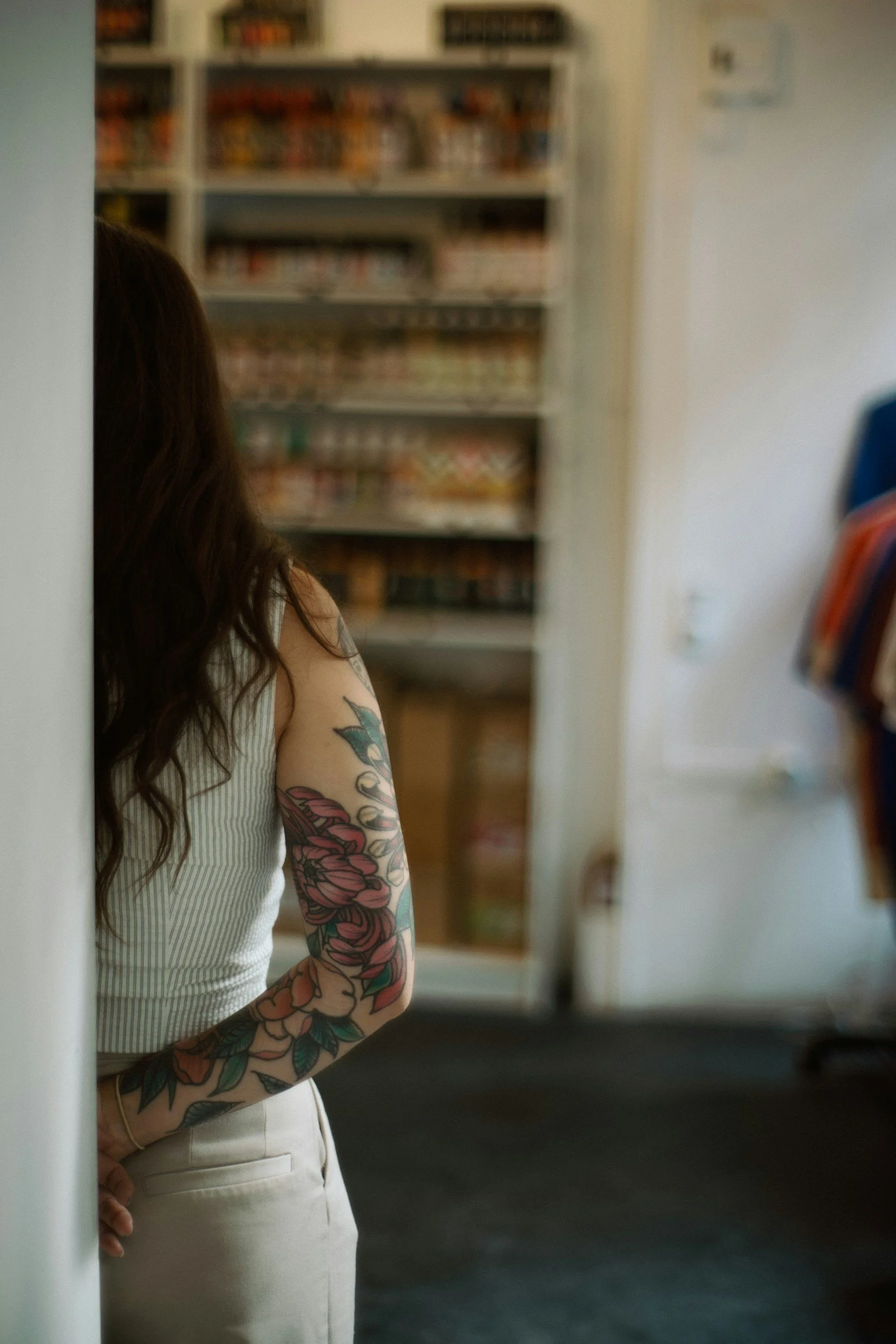 Partial view of a woman with long brown hair, wearing a sleeveless striped top, standing in front of shelves filled with various products in a store or library, with a tattooed arm visible.