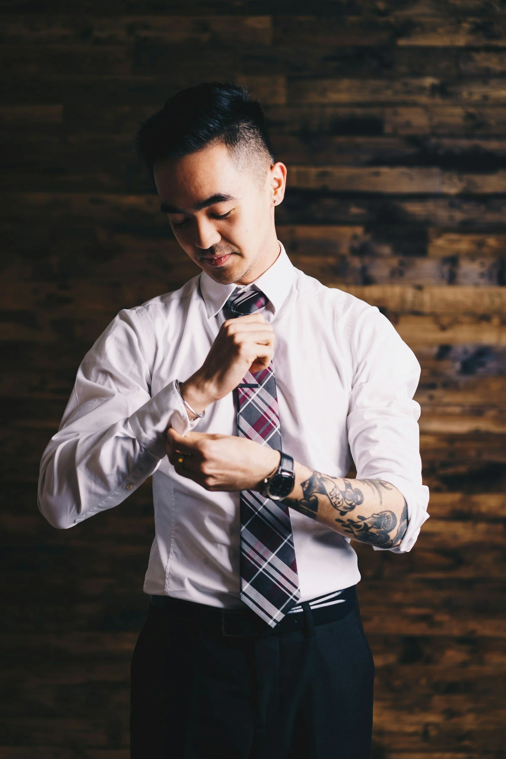 Man with tattoos on left arm adjusting his cufflink, wearing a white shirt, plaid tie, black pants, and a watch, standing in front of a wooden wall.