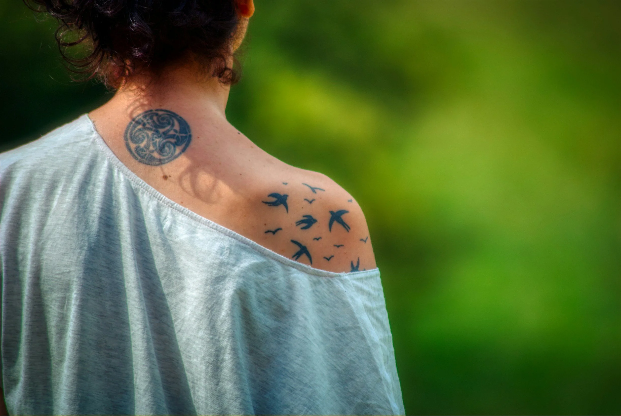 A person with short curly hair showing a large circular tattoo on the neck and a tattoo of flying birds on the shoulder, wearing a loose white shirt. The background is blurred green foliage.