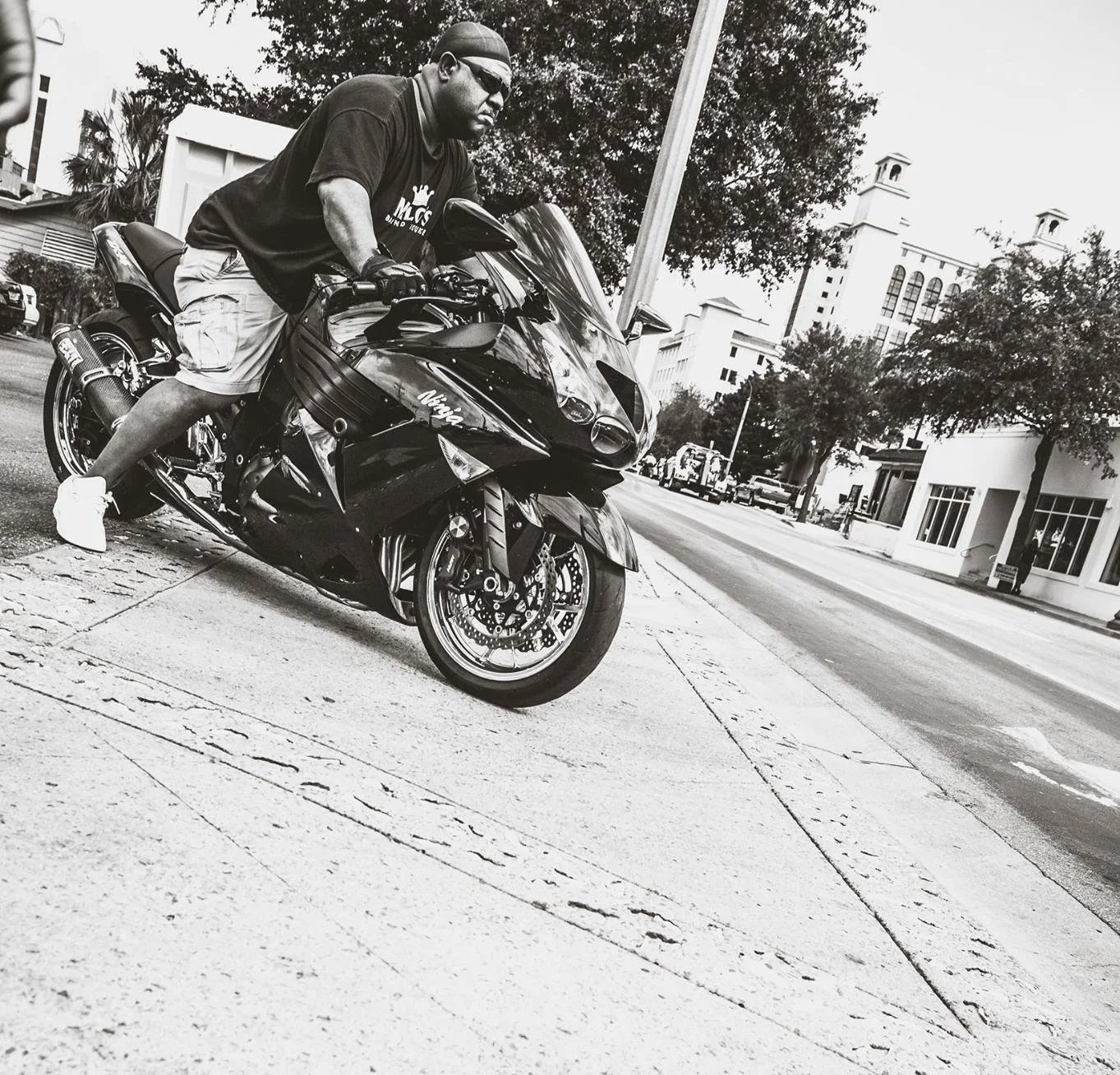 A man wearing sunglasses, a t-shirt, shorts, and sneakers pushes a black Kawasaki Ninja motorcycle on a city sidewalk in black and white.