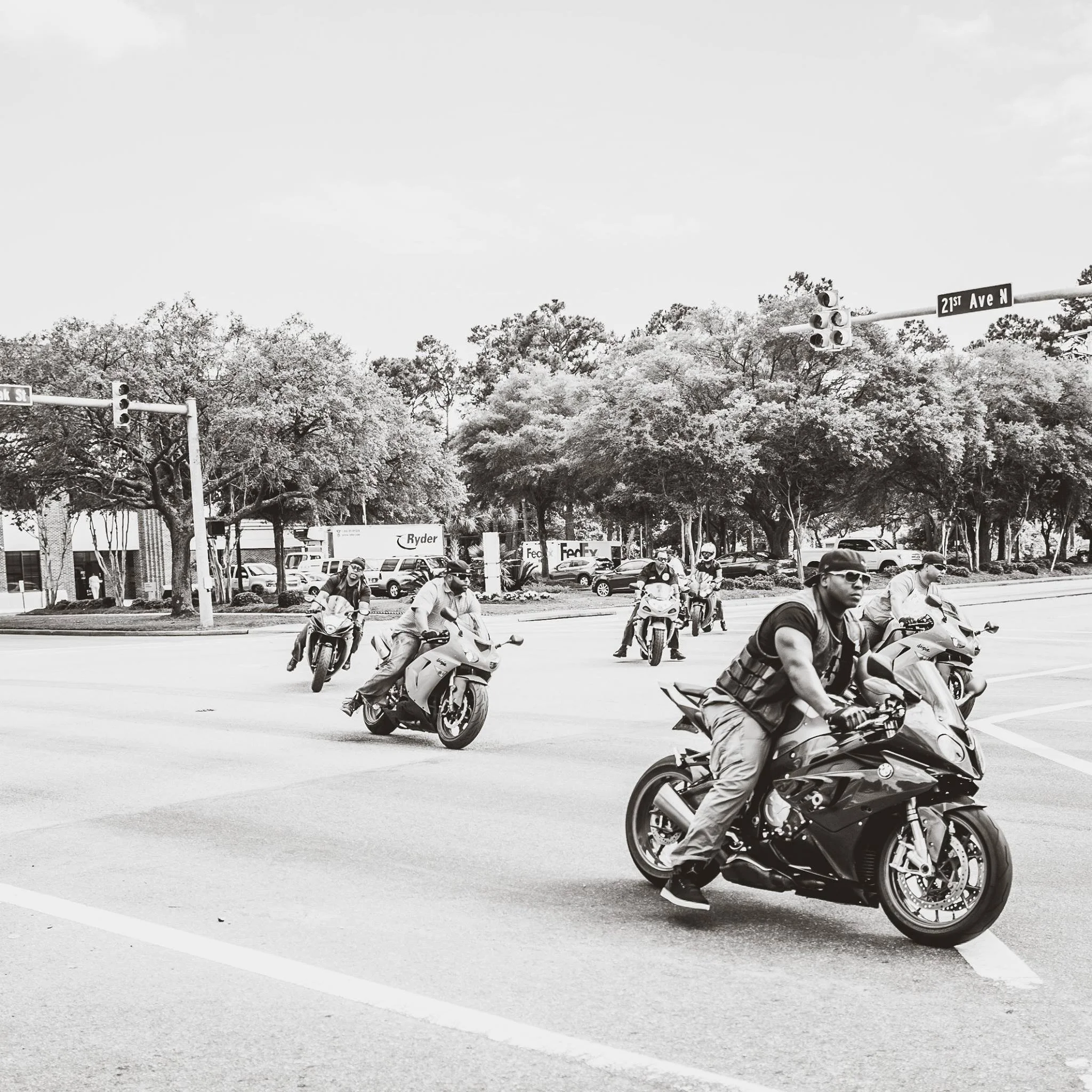 A group of motorcyclists riding through an intersection with traffic lights and street signs, trees, and parked cars in the background.