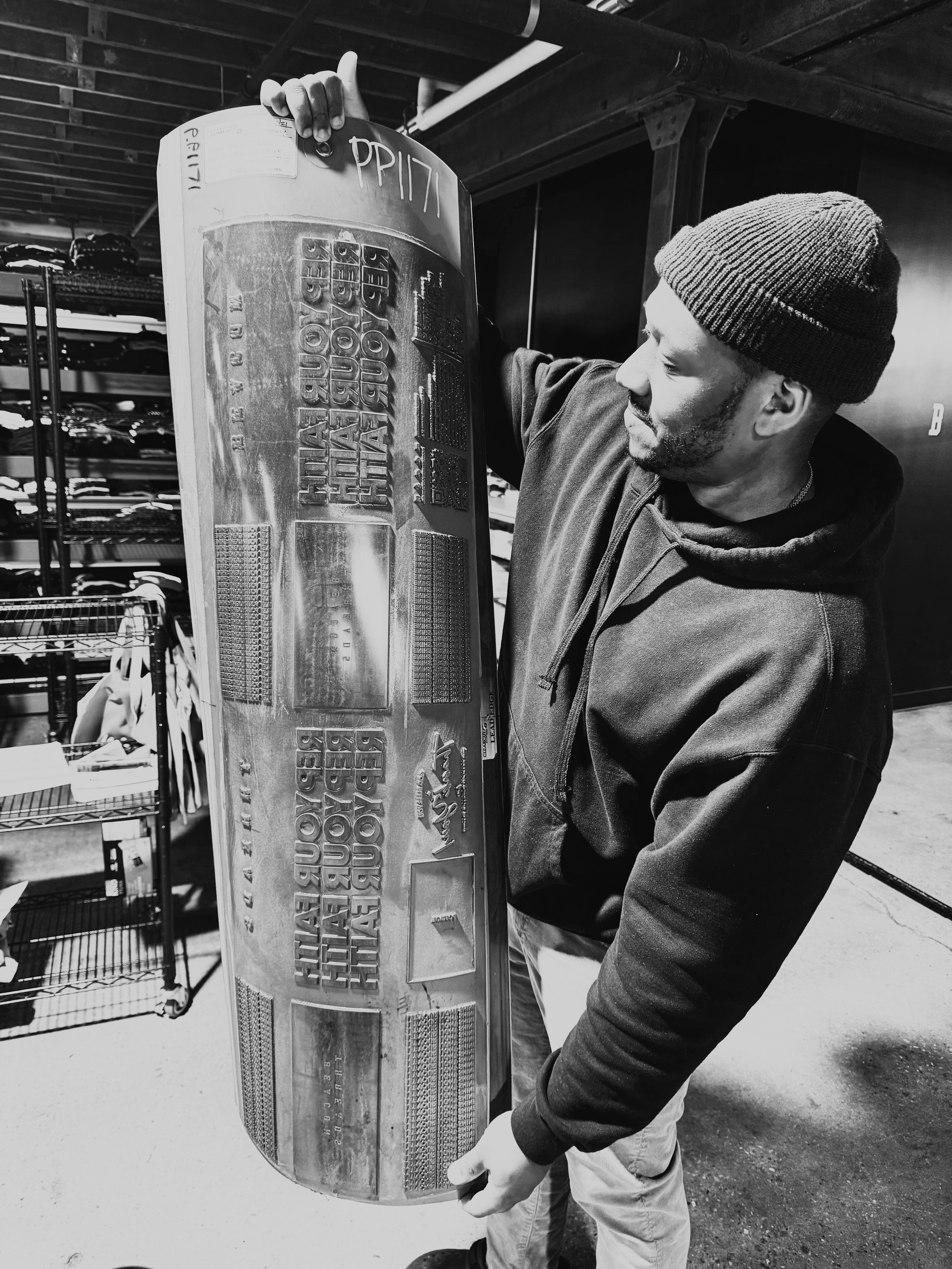 A man in a beanie and hoodie is holding up a skateboard with both hands, inspecting it. The skateboard is decorated with text and graphics, and the background shows shelves with clothing.