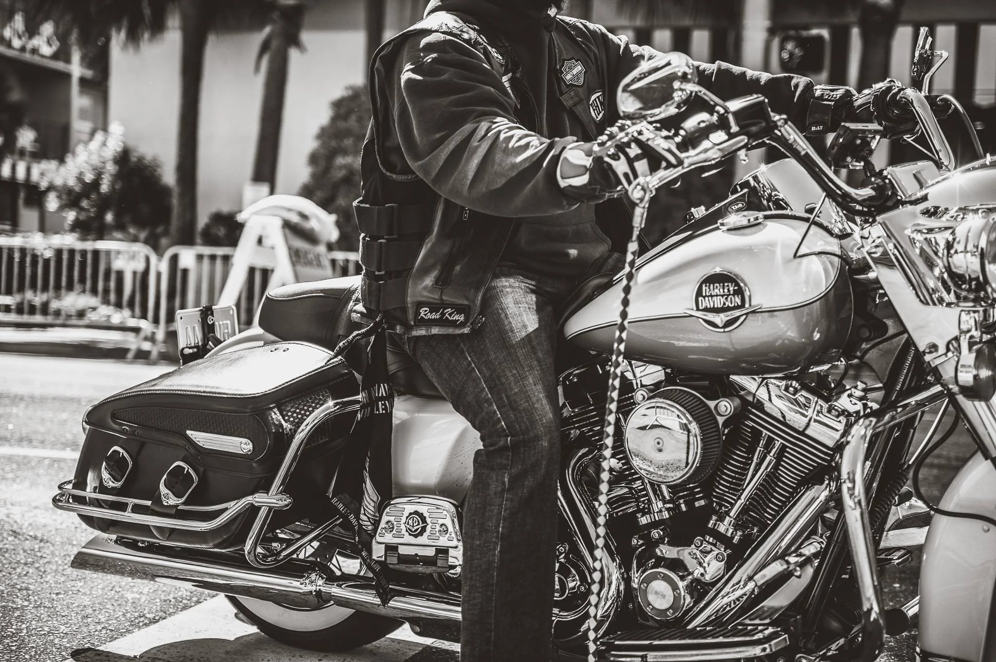 Black and white photo of a person sitting on a Harley-Davidson motorcycle, wearing a leather vest with patches, jeans, and gloves, with trees and a fence in the background.