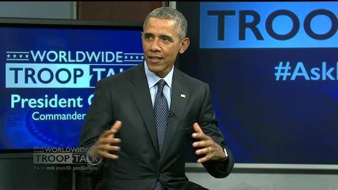 Former President Barack Obama speaking during a televised event, with a screen behind him displaying 'Worldwide Troop Talk' and hashtags.
