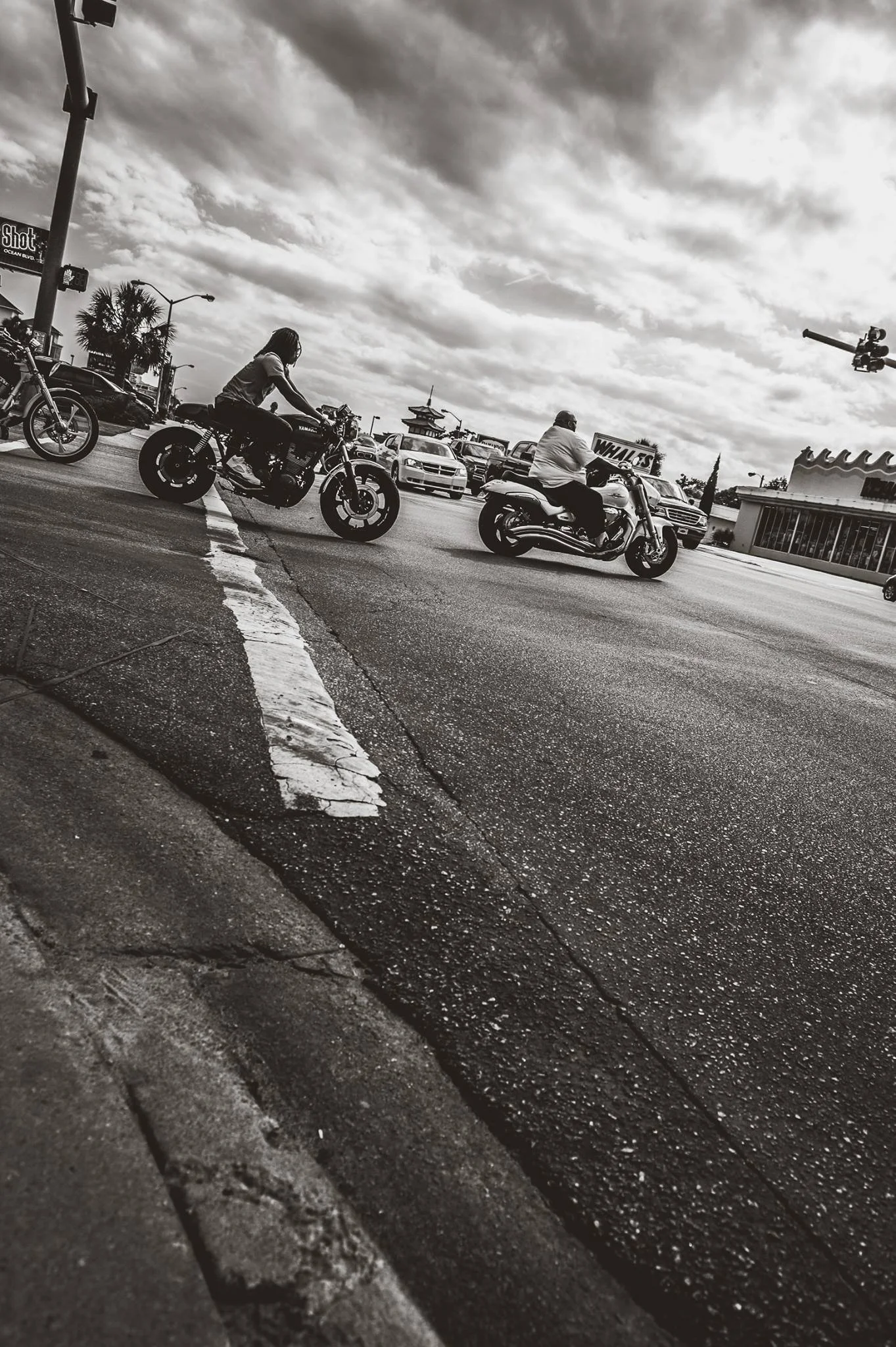Black and white photo of a street corner with two motorcycle riders crossing the intersection; one rider with long hair on a cruiser motorcycle, and the other with a cap on a larger motorcycle. Several cars are parked along the street, and a building