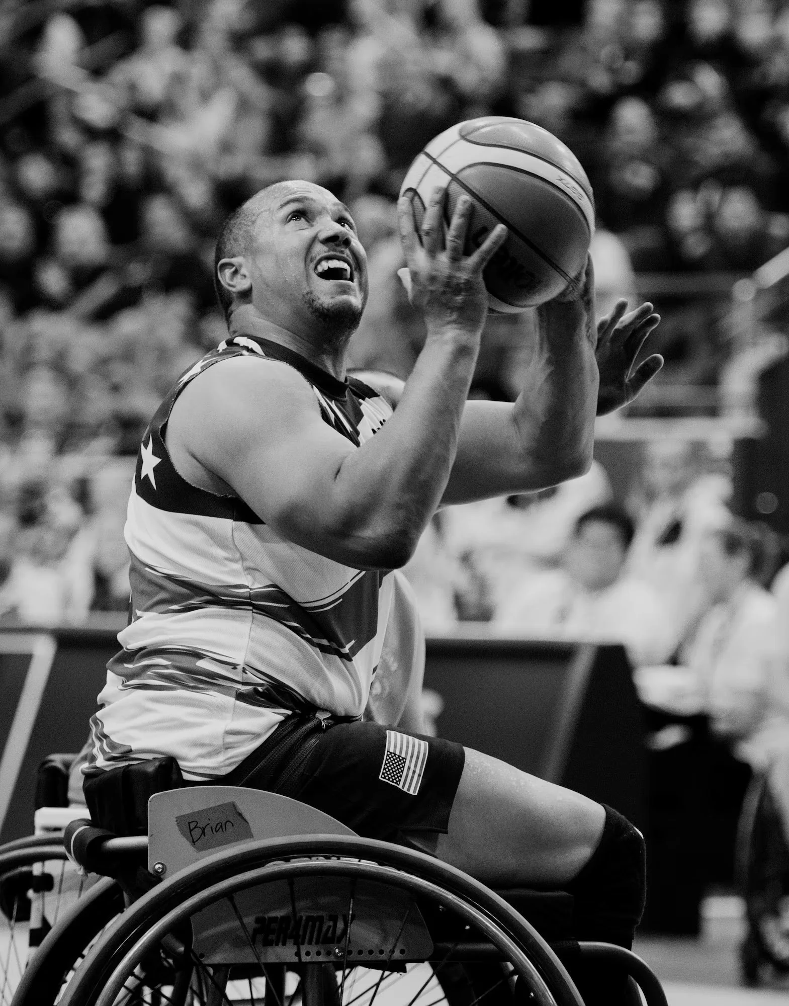 A male wheelchair basketball player in a sports uniform holding a basketball above his head during a game, with a focused expression. The crowd is blurred in the background.
