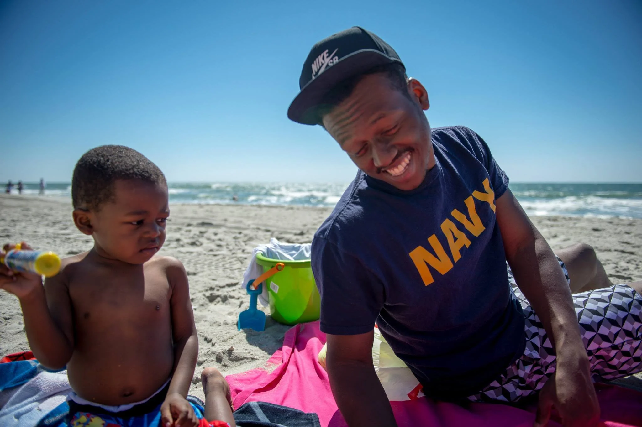 A young man and a small child sitting on the sandy beach, smiling and playing with beach toys, with the ocean in the background on a sunny day.