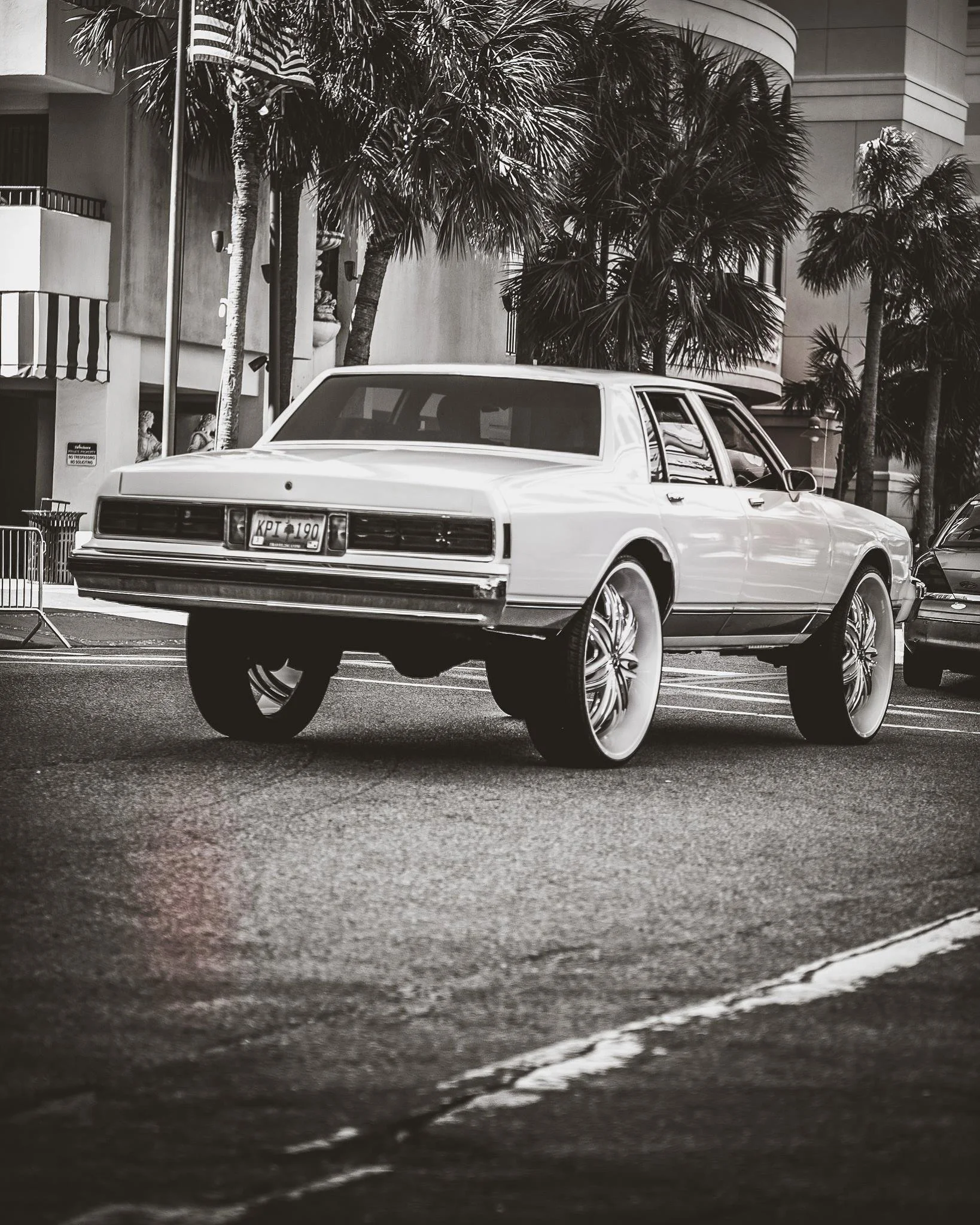 A vintage car with oversized wheels parked on the street with palm trees and buildings in the background, black and white photograph.