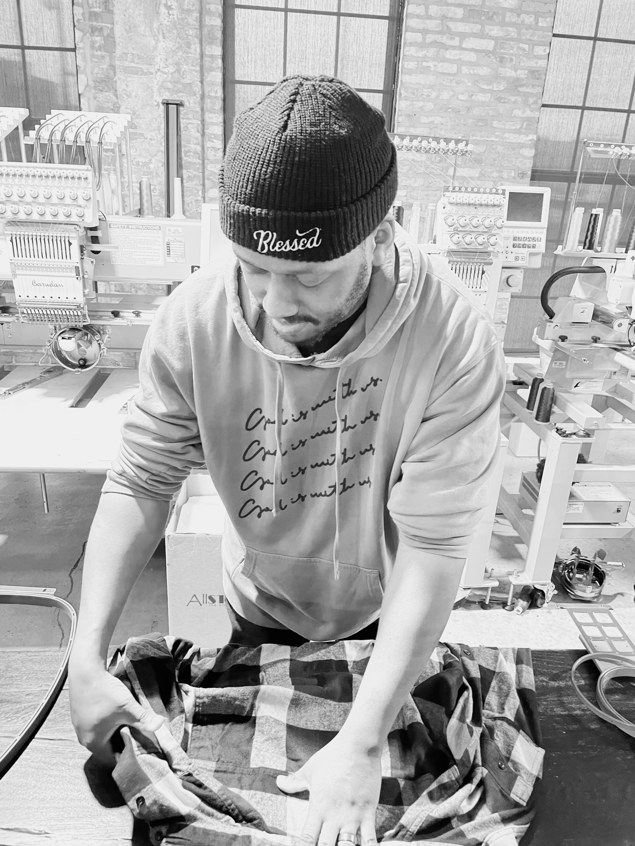A man wearing a beanie with the word 'Blessed' and a hoodie is folding a checkered shirt on a table in a workshop with embroidery machines in the background.
