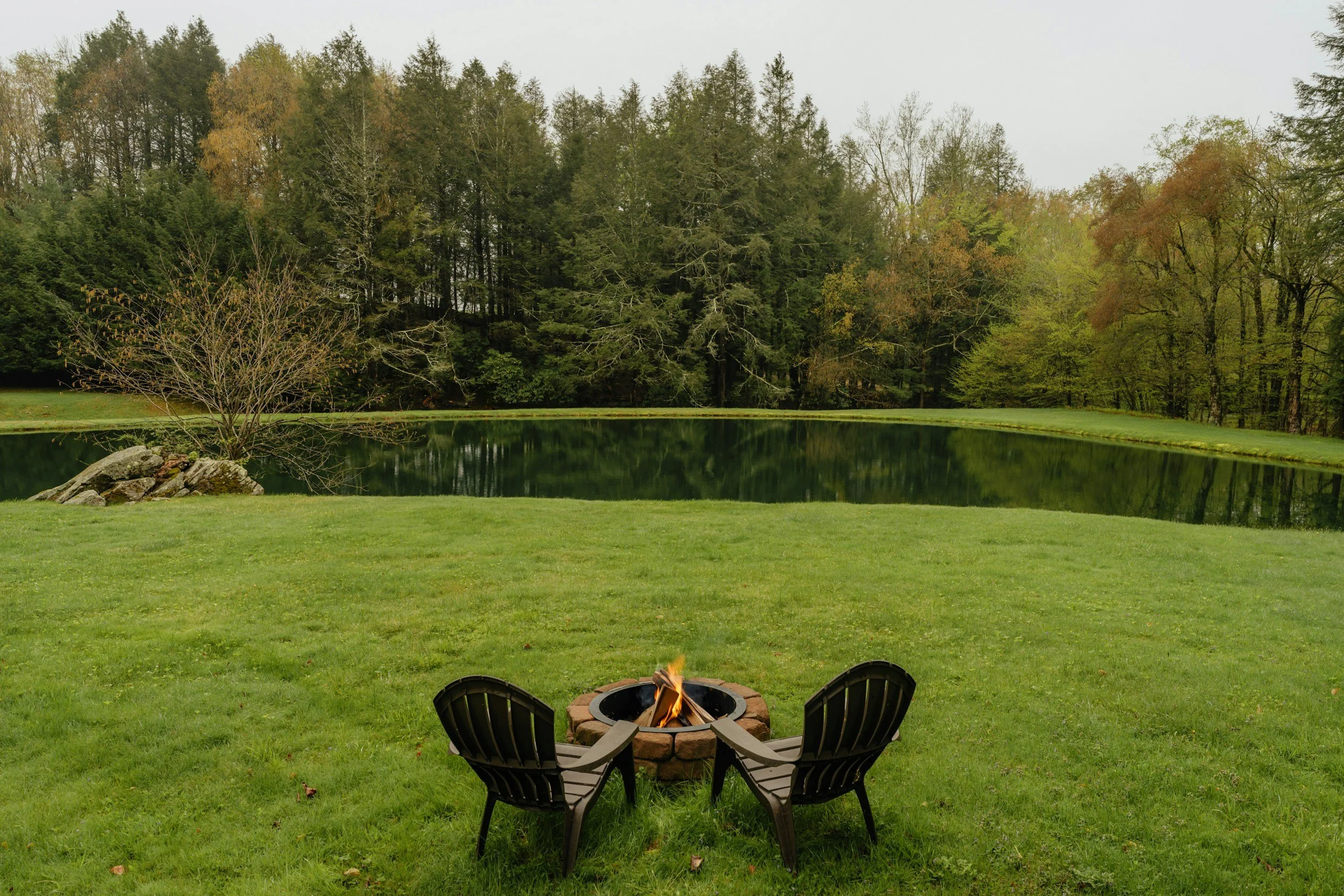 A backyard with two chairs surrounding a small fire pit on a grass lawn, overlooking a pond with trees in the background.