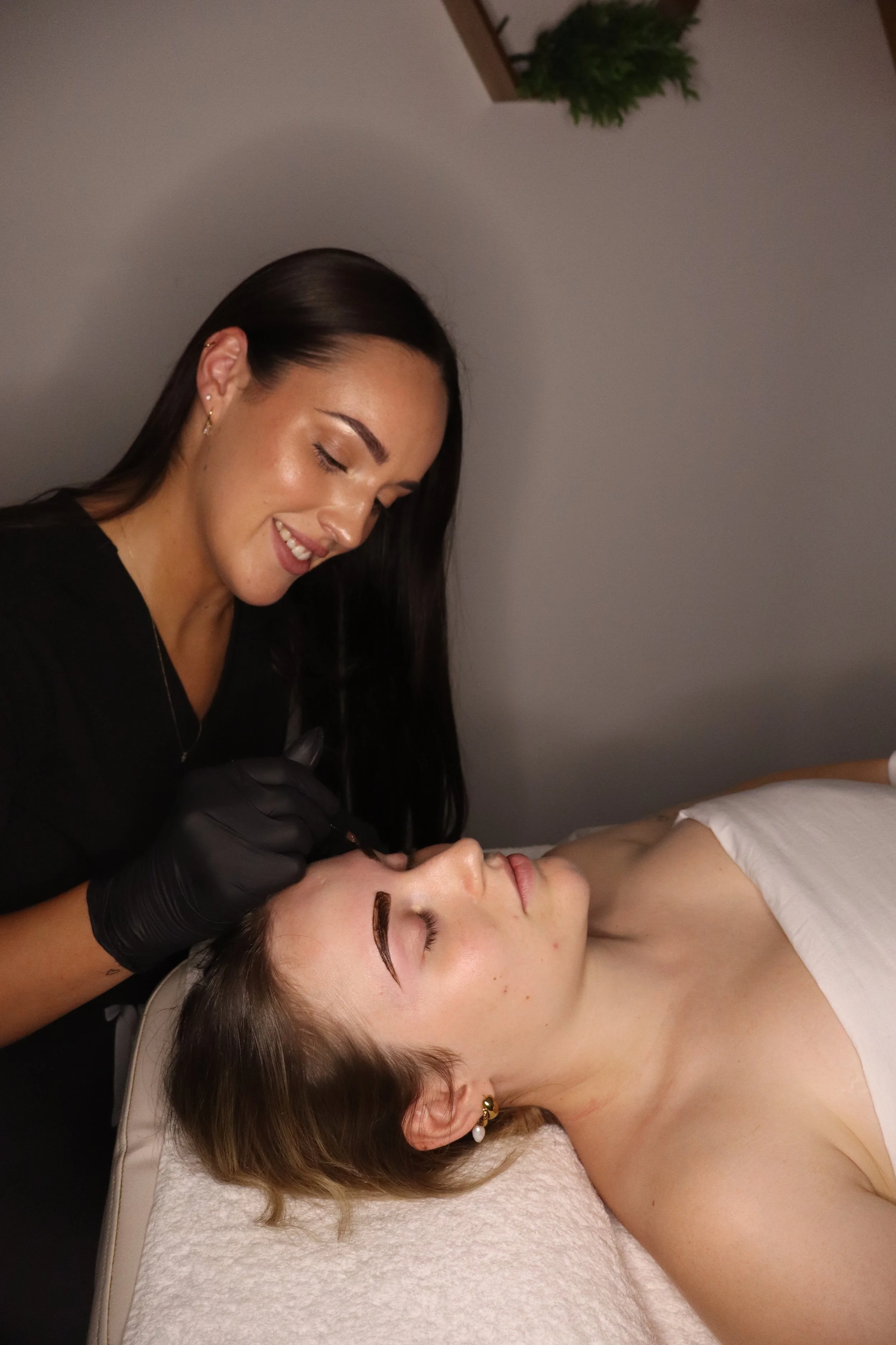 A woman lying on a bed receiving a cosmetic eyebrow tattooing procedure from an artist wearing black gloves.