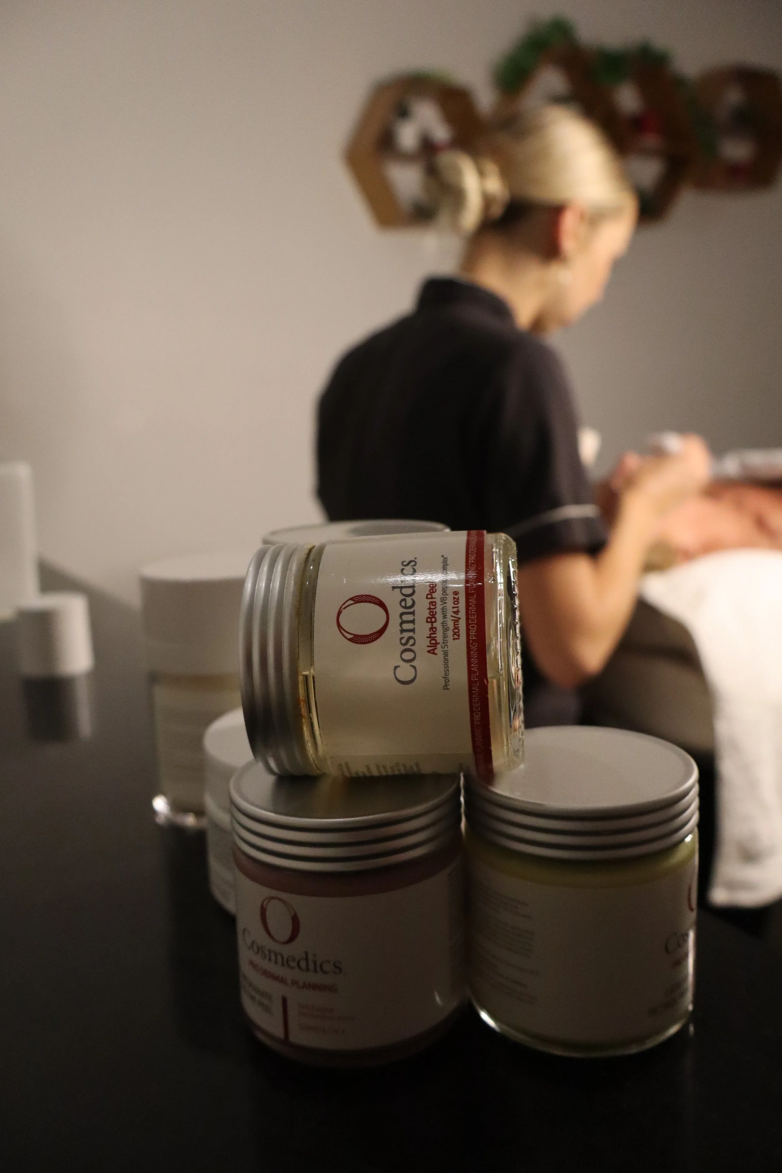 Several containers of Cosmedics skin care product on a black surface, with a woman in a dark uniform blurred in the background.