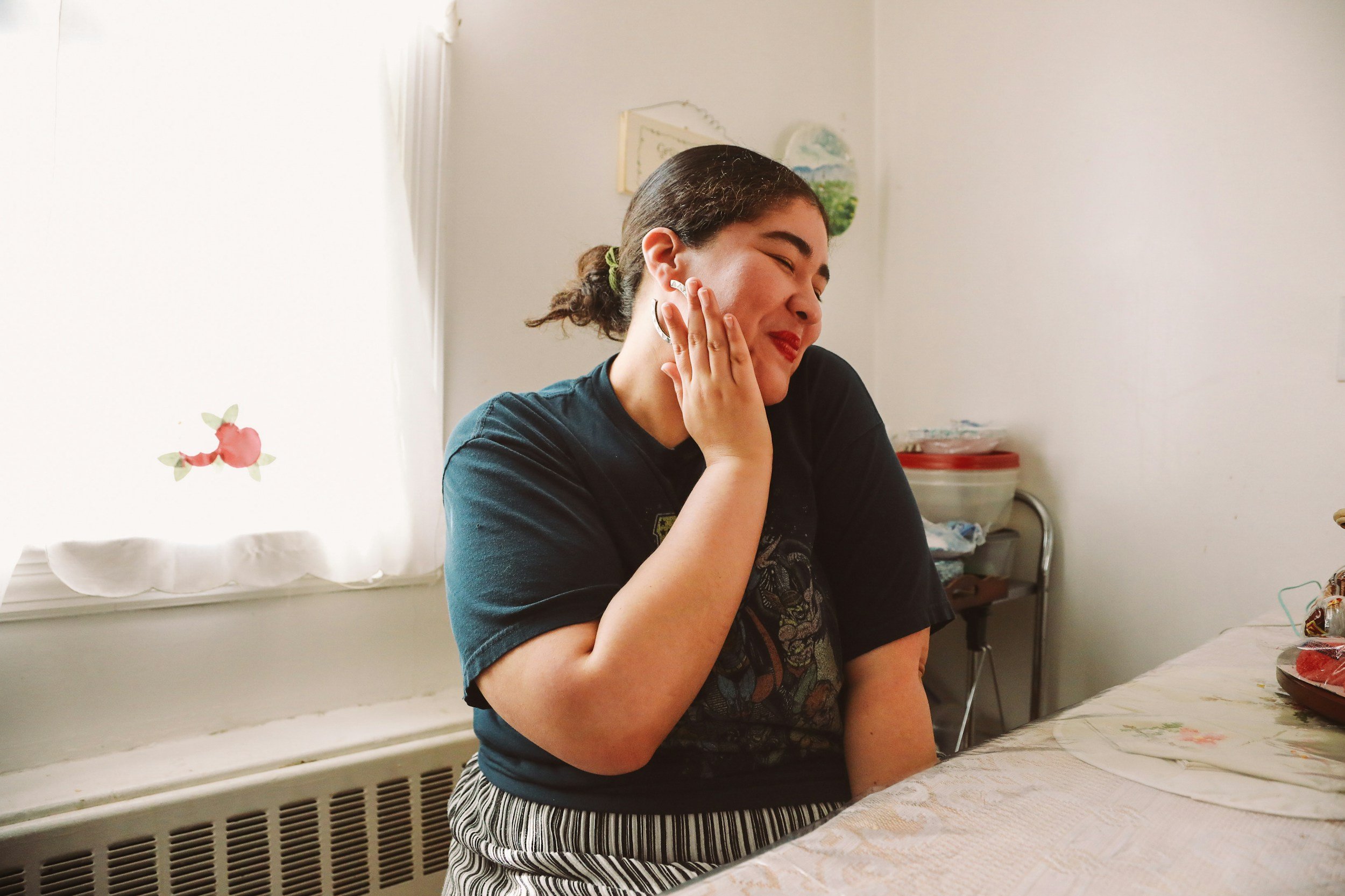 A young woman sitting at a table, smiling and touching her face with her eyes closed, in a cozy room with white walls and a window with curtains.