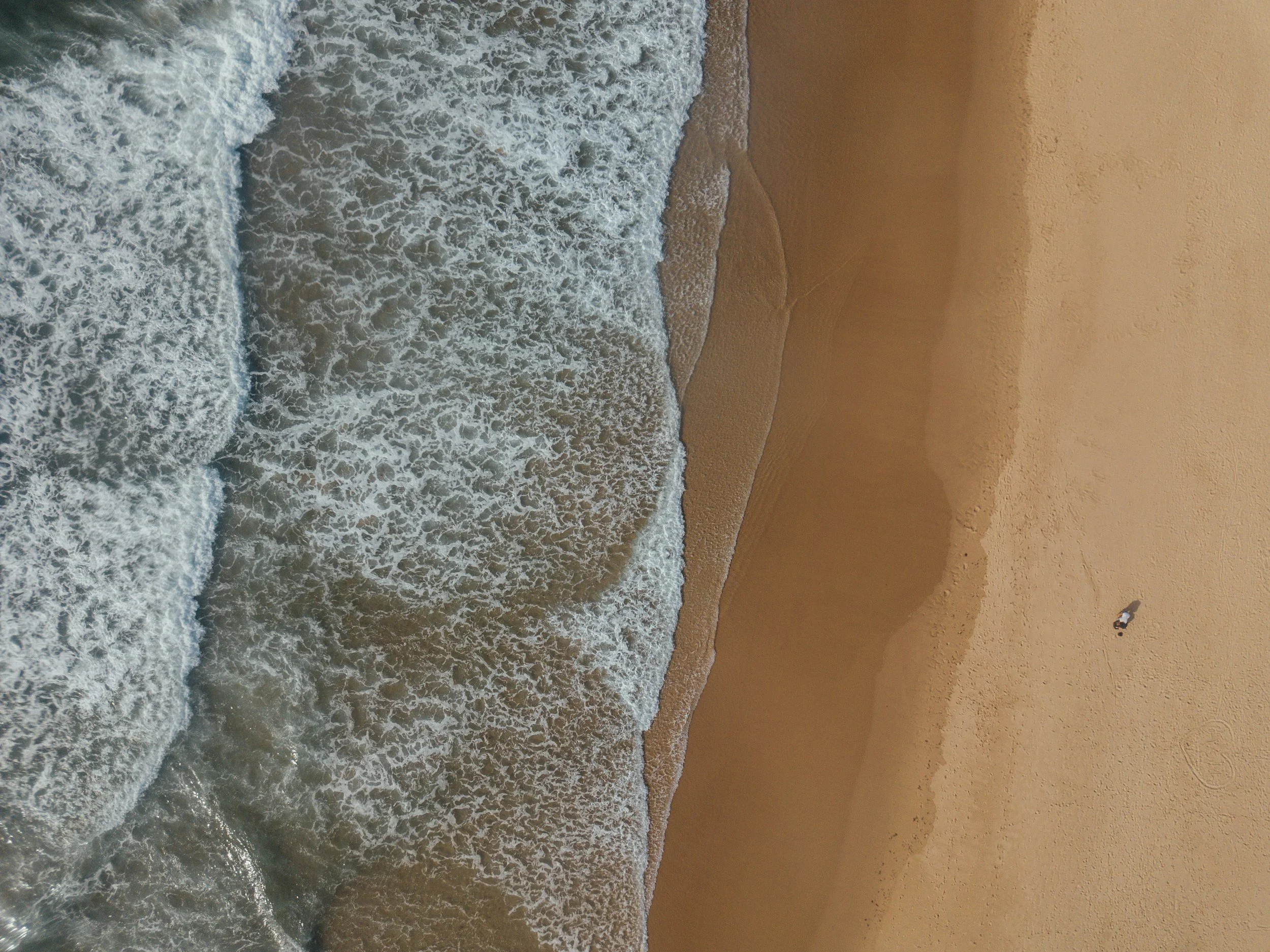 Aerial view of ocean waves crashing onto sandy beach with a person walking along the shoreline.