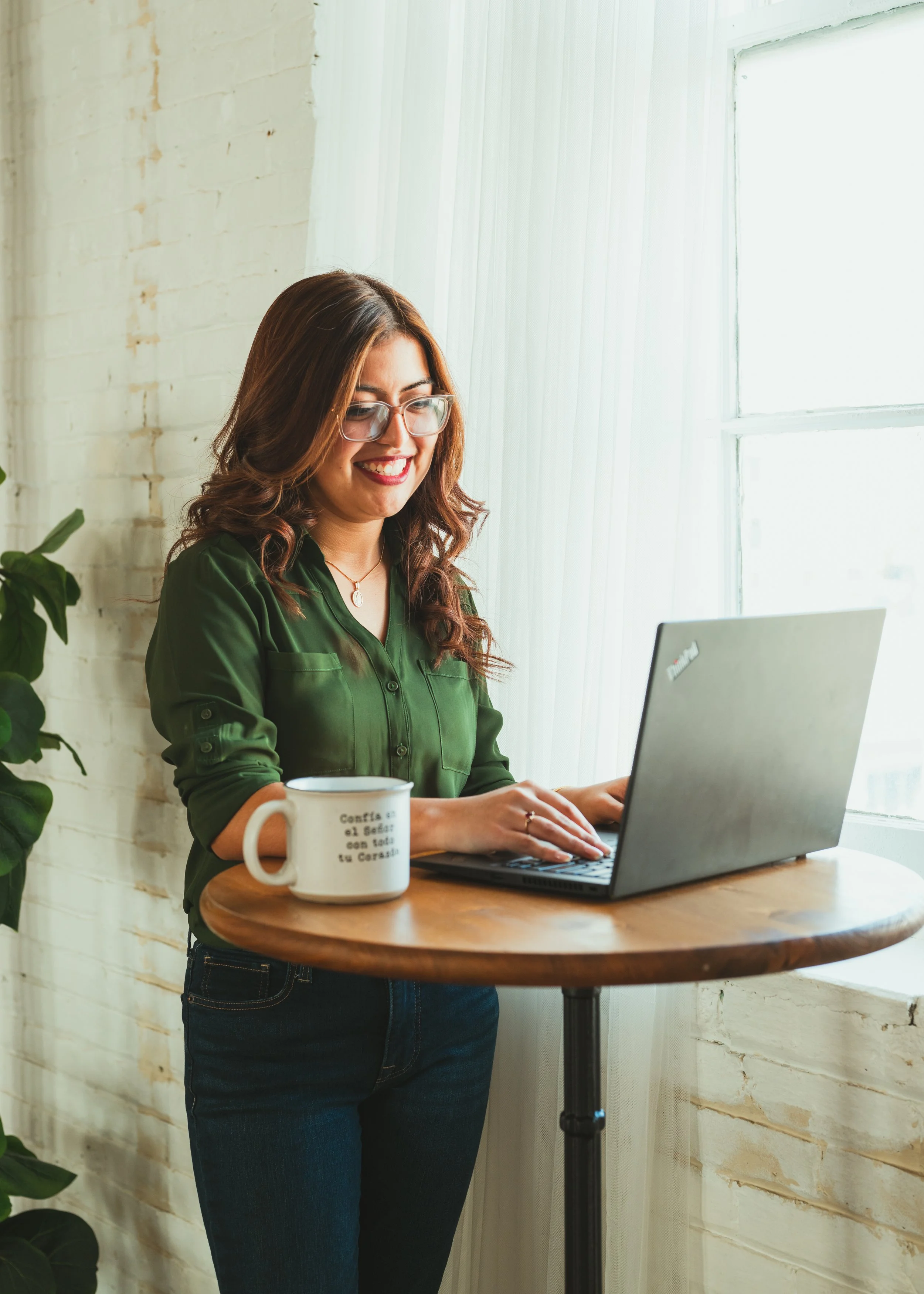Latina bilingual therapist smiling while using a laptop for virtual therapy sessions, white mug nearby, bright natural light through large windows.