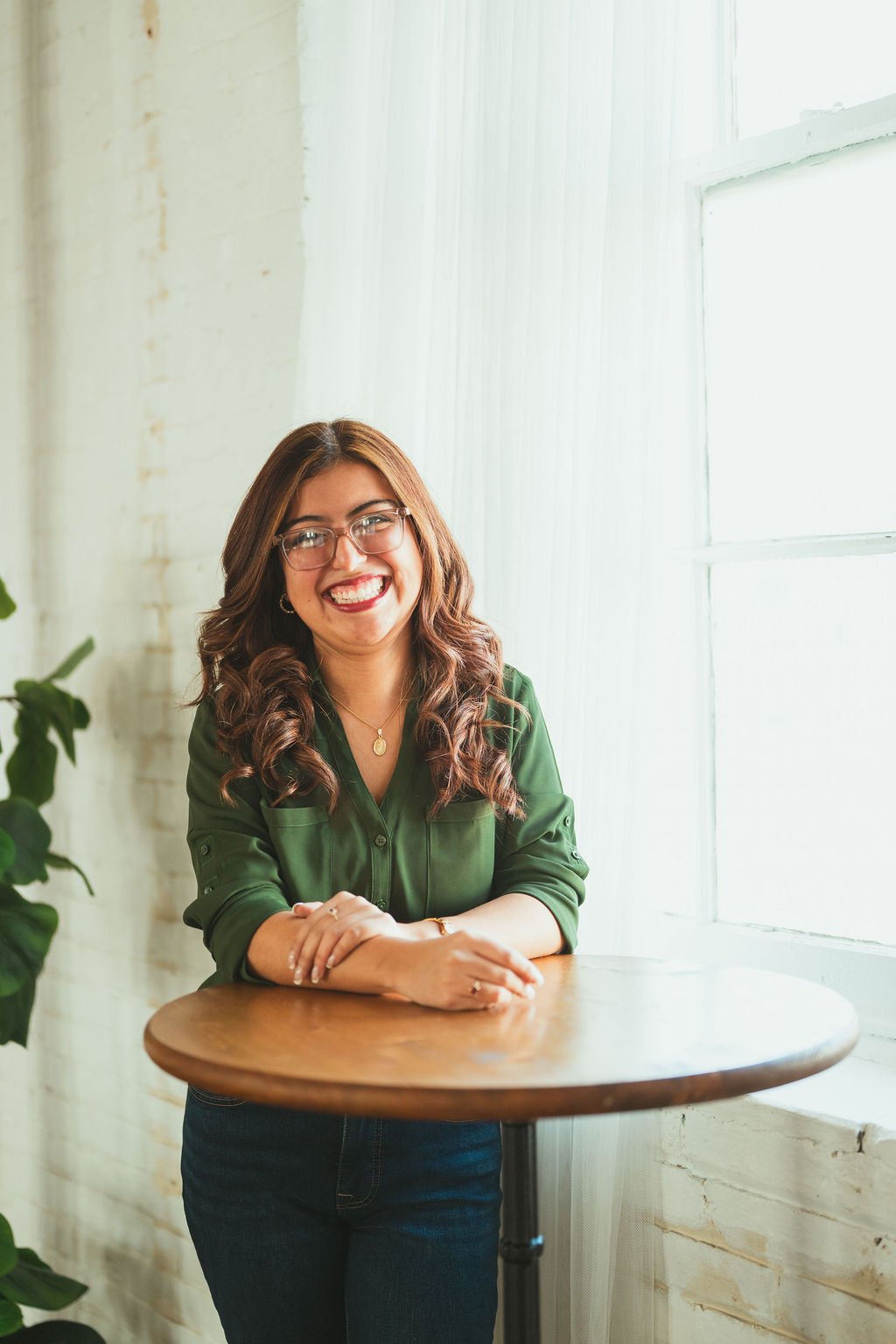 1. Latina bilingual therapist smiling beside a large window in a cozy, plant-filled room, appearing warm and approachable.