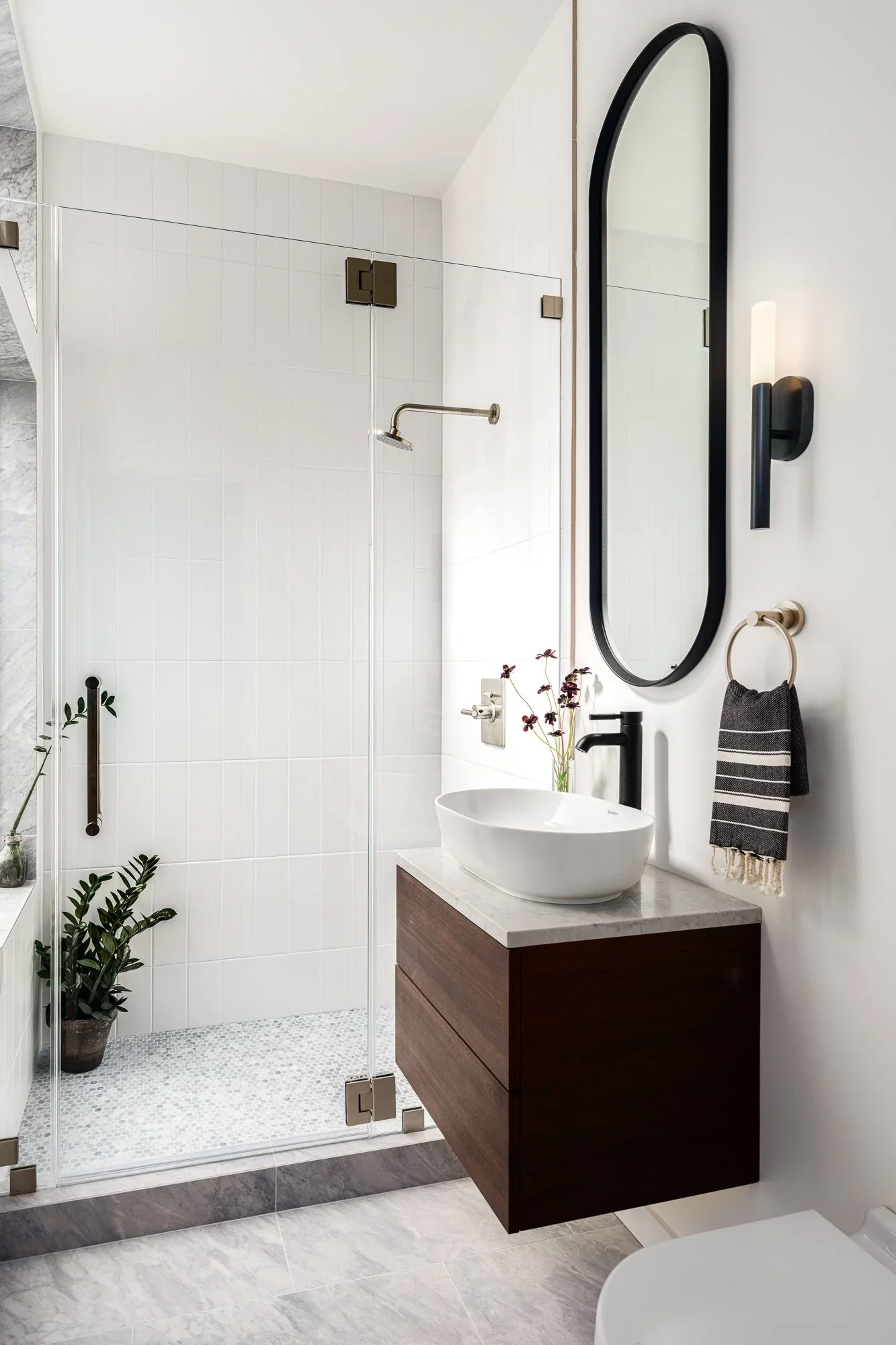 Floating dark walnut vanity with white vessel sink, marble countertop, black oval mirror, matte black sconce, and frameless glass shower with white vertical tile — Pacific Heights San Francisco residence by Mokume Design Studio