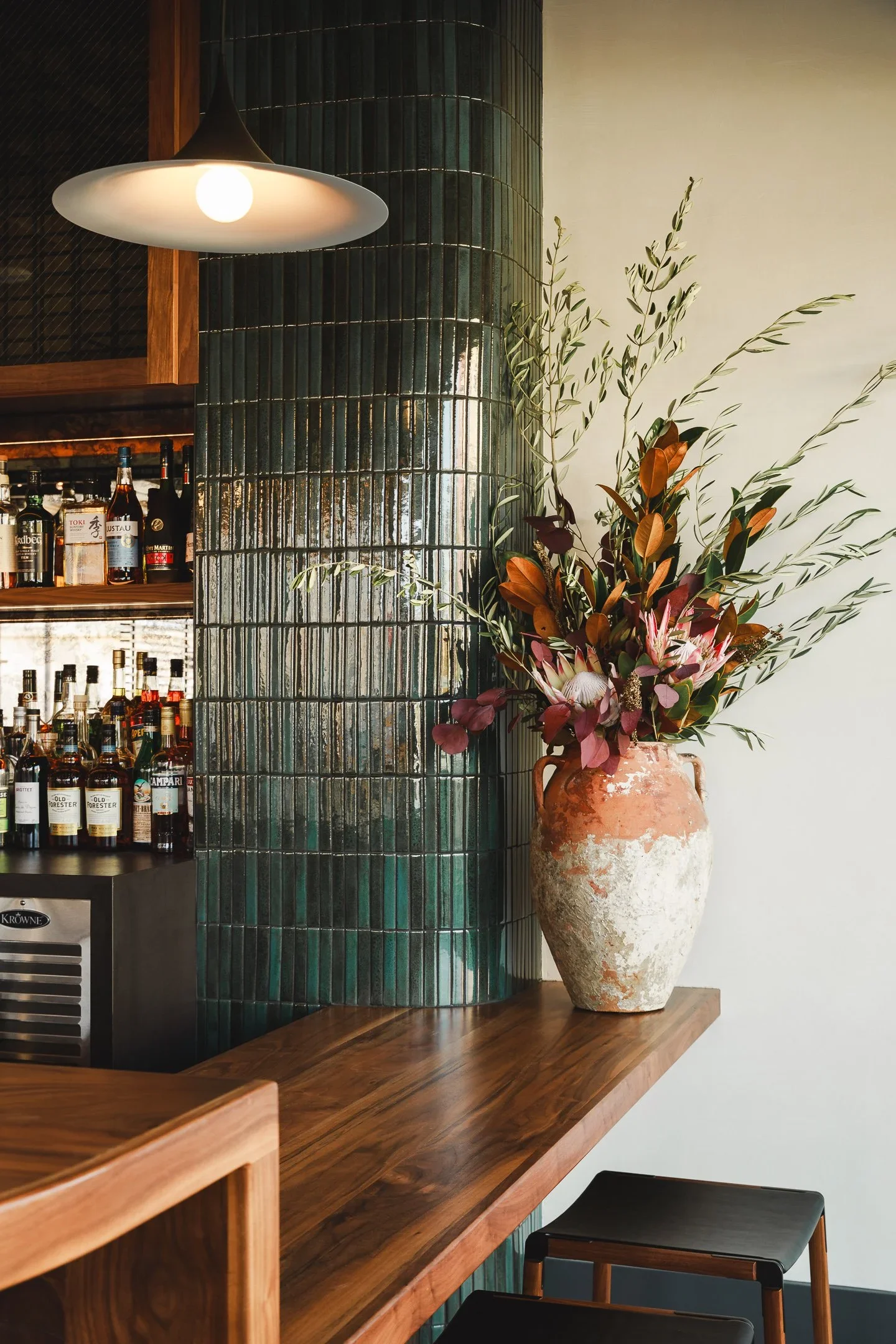 Warm walnut bar with full liquor display, antique mirror shelving, wood and black leather barstools, and terrazzo tile floor — Marlena restaurant bar design Long Beach by Mokume Design Studio