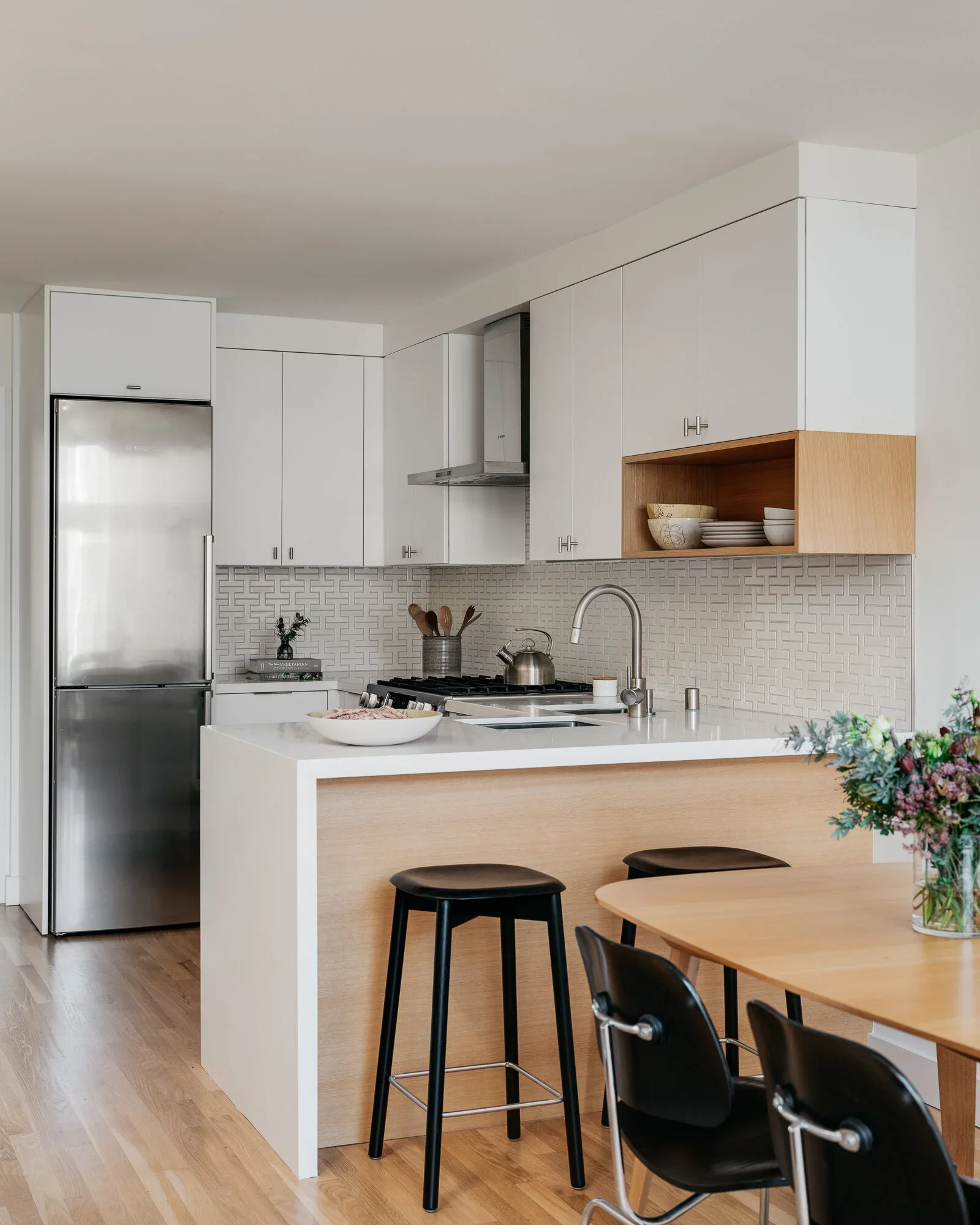 Compact white flat-front kitchen with natural oak peninsula, geometric tile backsplash, black barstools, and open oak display shelf — Nopa Loft San Francisco by Mokume Design Studio