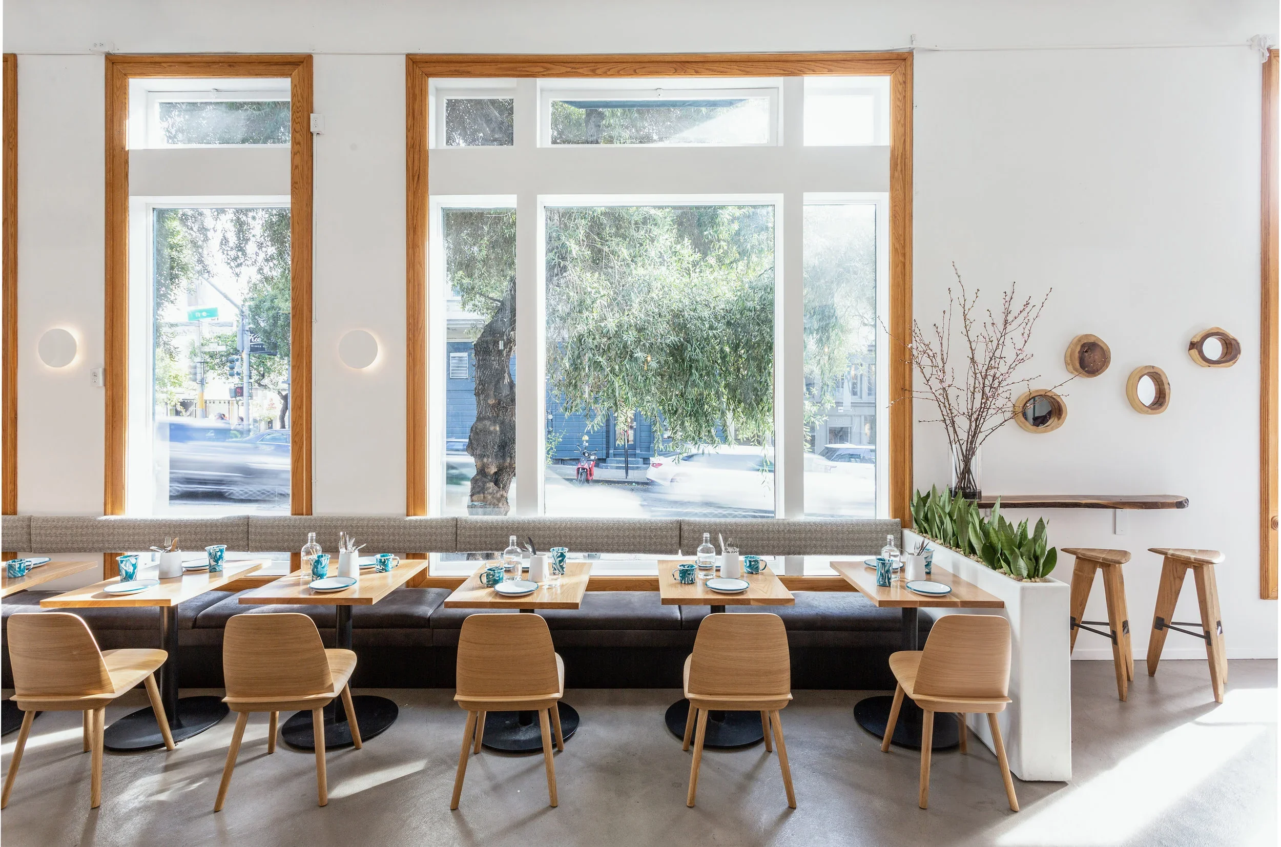 Noosh restaurant dining room with floor-to-ceiling oak-framed windows, gray upholstered banquette, natural oak chairs, and blue marbled ceramic place settings — San Francisco restaurant interior by Mokume Design Studio