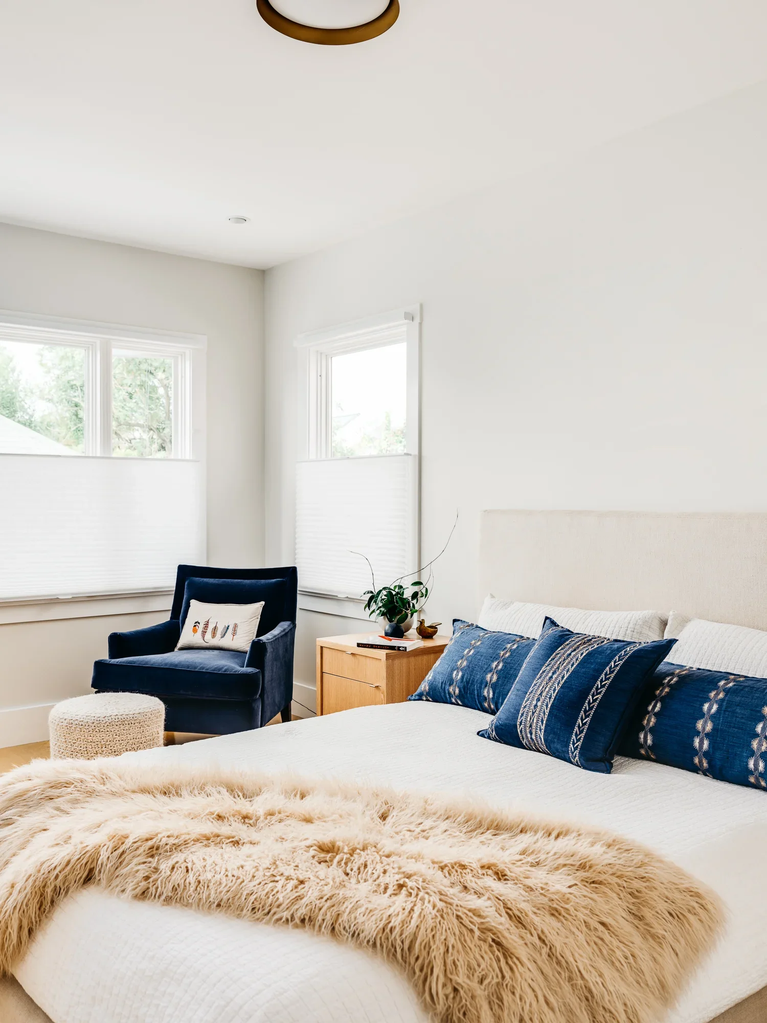 Bright primary bedroom with upholstered linen headboard, navy blue velvet accent chair, layered indigo throw pillows, and shearling blanket — modern residential interior design by Mokume Design Studio