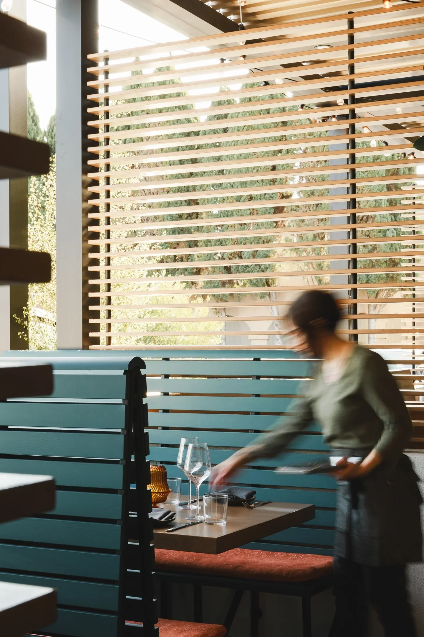Server placing glassware on a sunlit dining table set with wine glasses, amber votives, and linen napkins — Marlena restaurant Long Beach by Mokume Design Studio