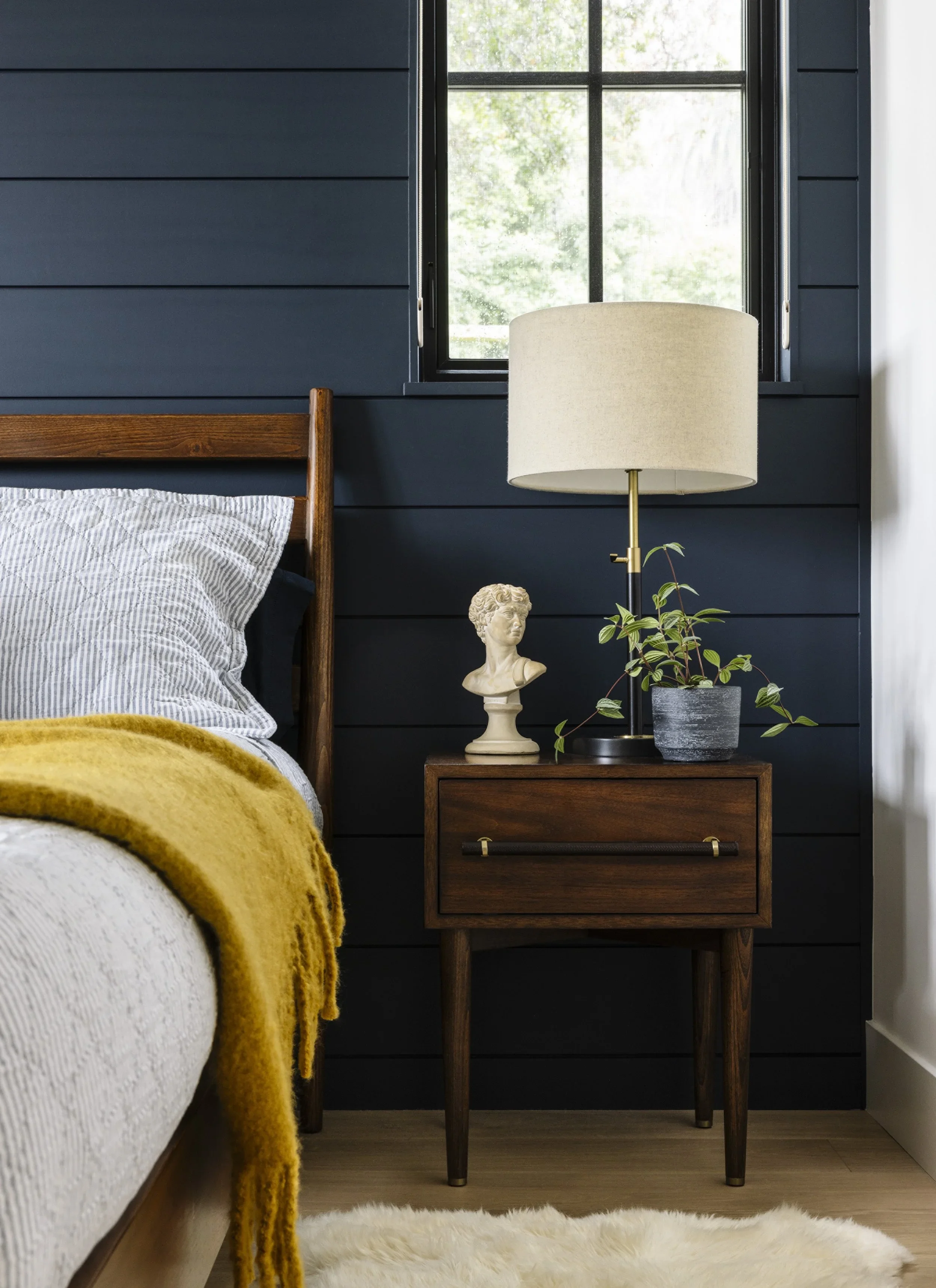 Close-up of walnut mid-century nightstand with brass lamp, classical bust sculpture, and trailing plant against navy painted shiplap wall — Corte Madera Residence by Mokume Design Studio