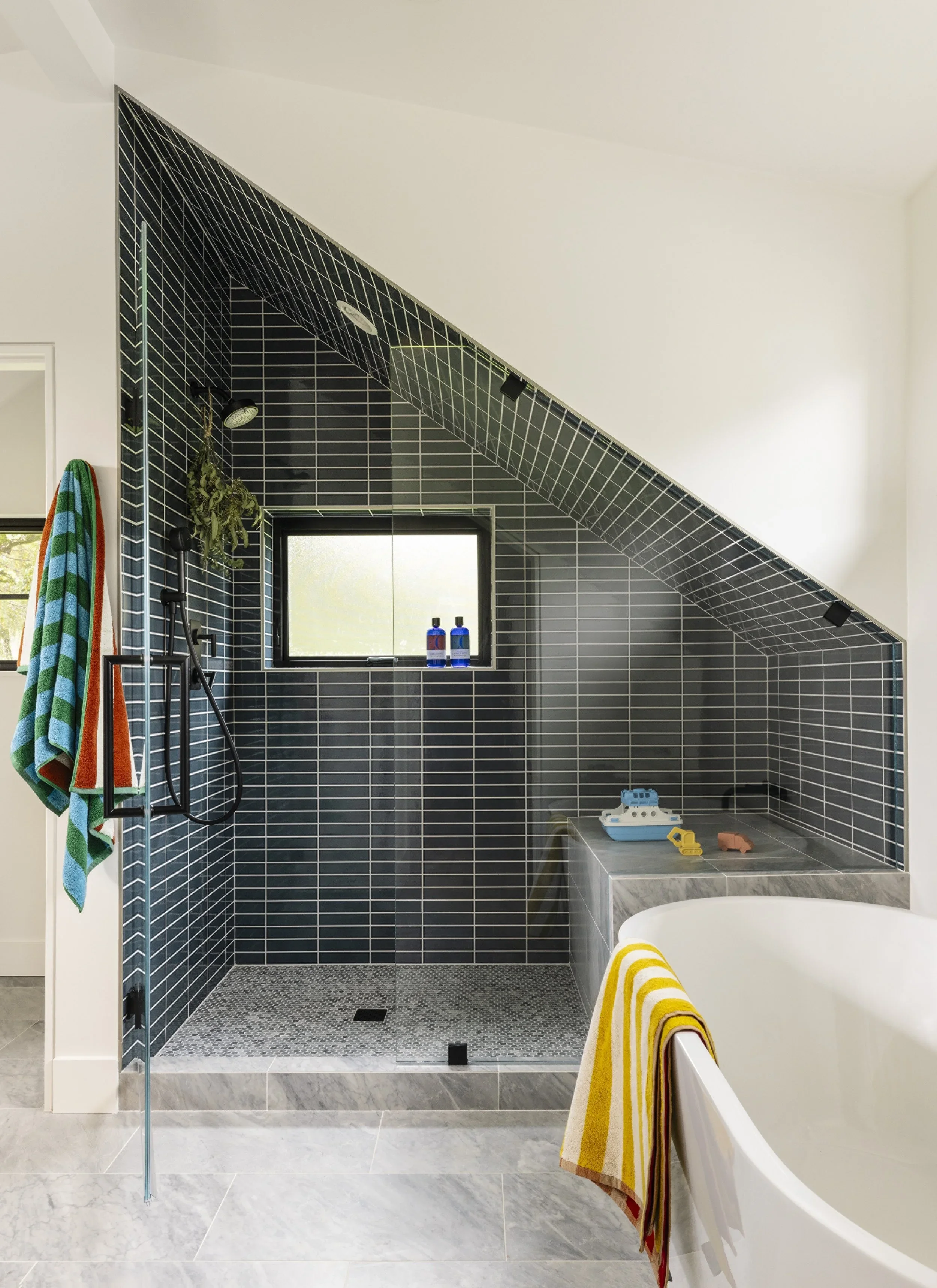 Children's bathroom with dramatic angled dark teal tile shower following roofline, freestanding soaking tub, marble floor tile, and colorful striped towels — Corte Madera Residence by Mokume Design Studio