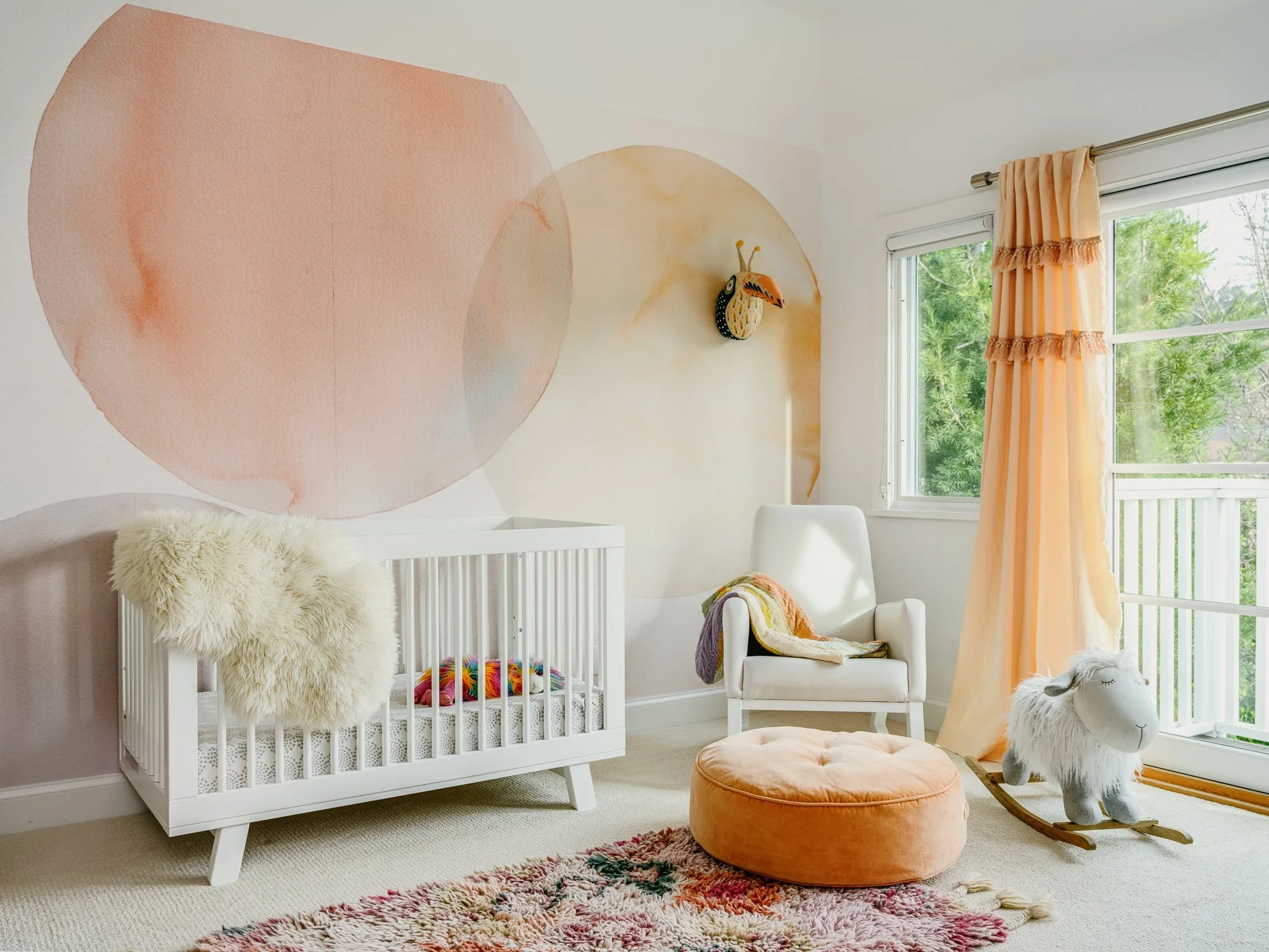 Bright nursery with large watercolor circle mural in blush and peach, white crib, velvet pouf, fringe curtains, and Moroccan shag rug — Kentfield California children's room design by Mokume Design Studio