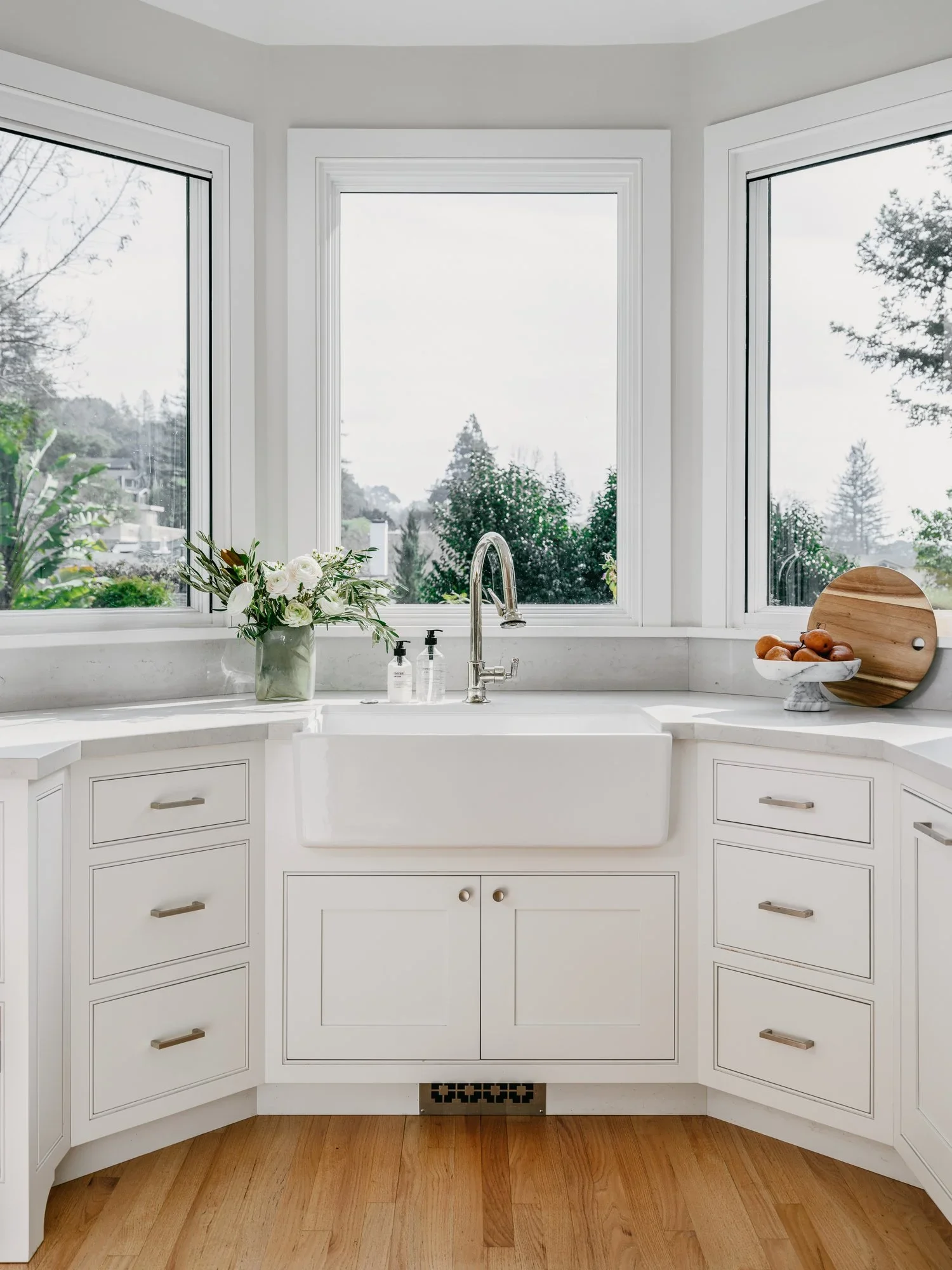 White farmhouse sink set into a curved bay window with panoramic garden views, white quartz countertop, and shaker cabinetry with brushed nickel hardware — Kentfield California kitchen remodel by Mokume Design Studio