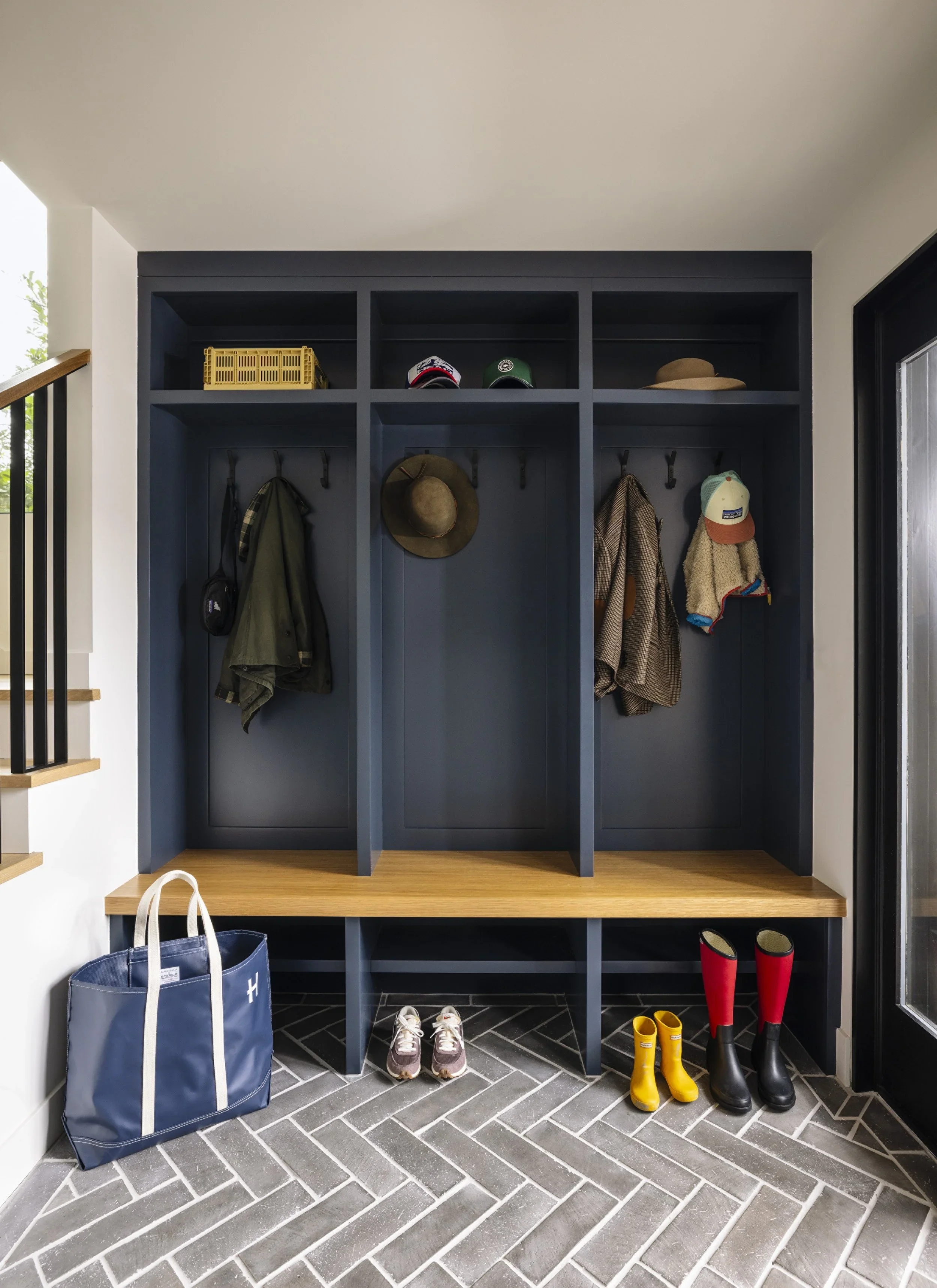 
Navy blue built-in mudroom lockers with individual coat hooks, upper shelving, natural oak bench, and herringbone slate tile floor — Corte Madera Residence by Mokume Design Studio