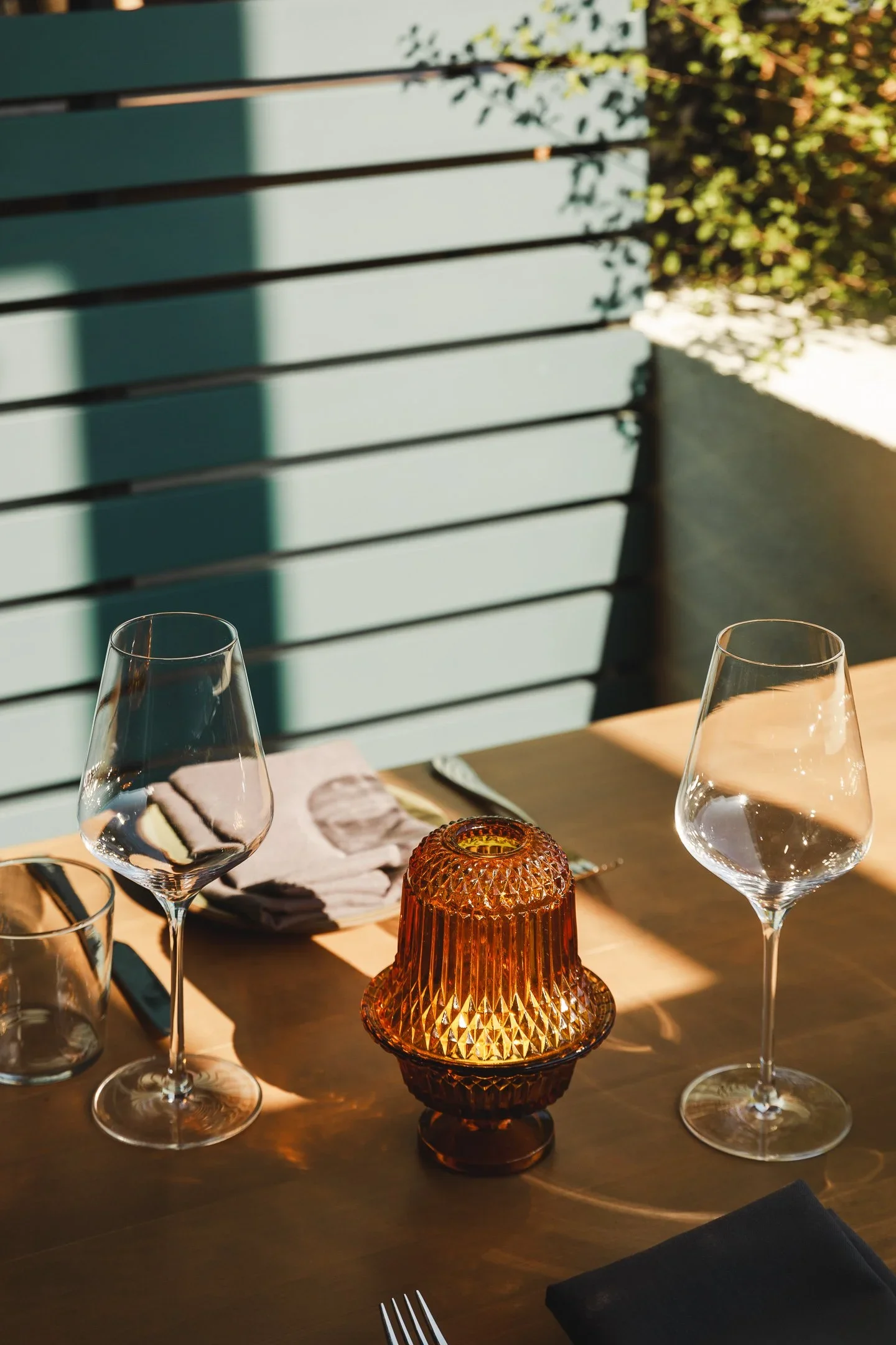 Sunlit outdoor dining table set with crystal wine glasses and amber glass votive candle against teal slatted privacy screen — Marlena restaurant Long Beach by Mokume Design Studio