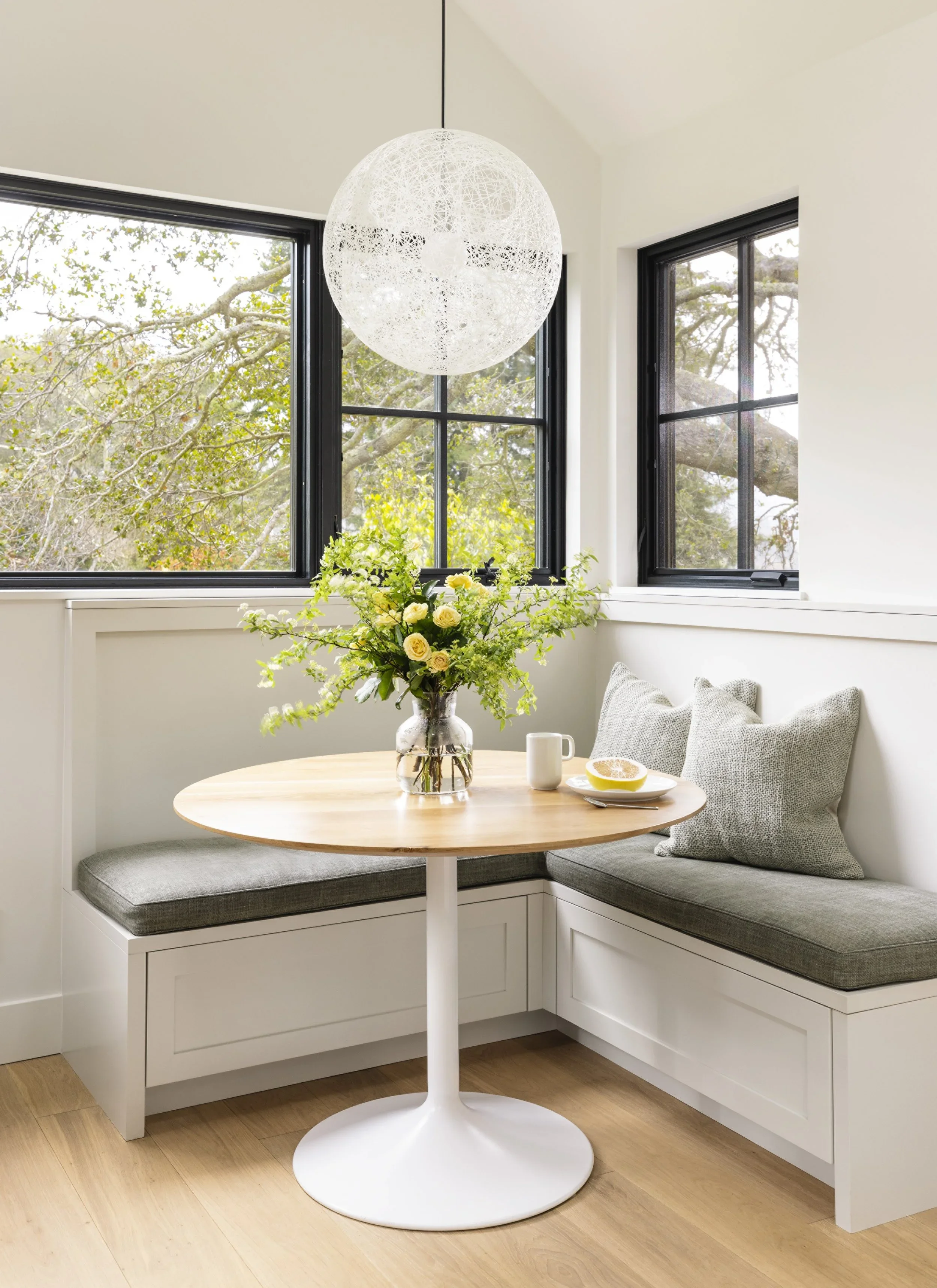 Built-in corner banquette with sage green cushions, round tulip pedestal table, globe pendant light, and black-framed windows overlooking garden — Corte Madera Residence by Mokume Design Studio
