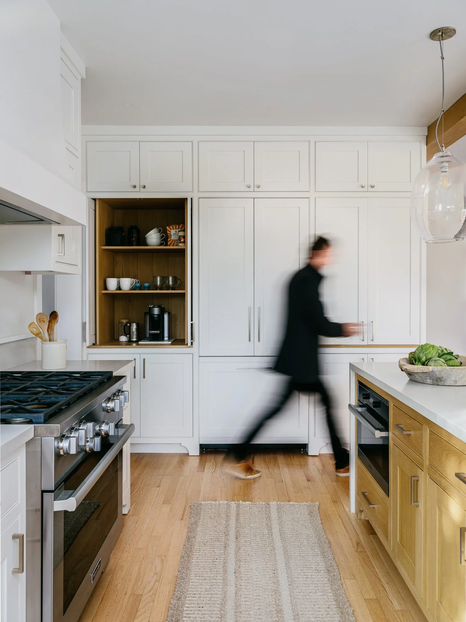 White shaker kitchen with open oak-lined coffee station cabinet, integrated appliances, and natural oak lower cabinetry with person in motion — Kentfield California home remodel by Mokume Design Studio