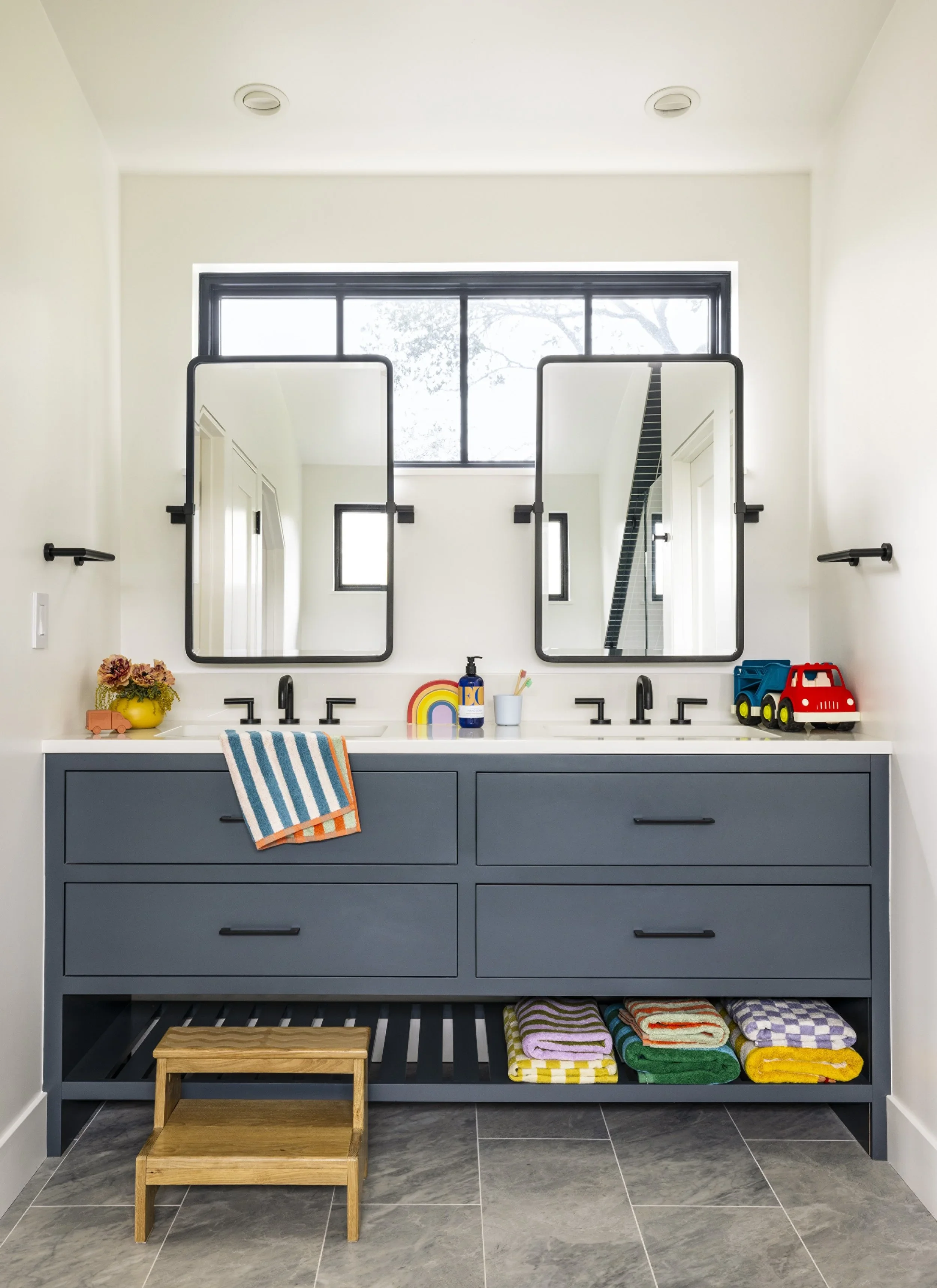 Children's double vanity with slate blue cabinetry, dual black-framed mirrors, matte black faucets, open lower shelf with colorful stacked towels, and wood step stool — Corte Madera Residence by Mokume Design Studio
