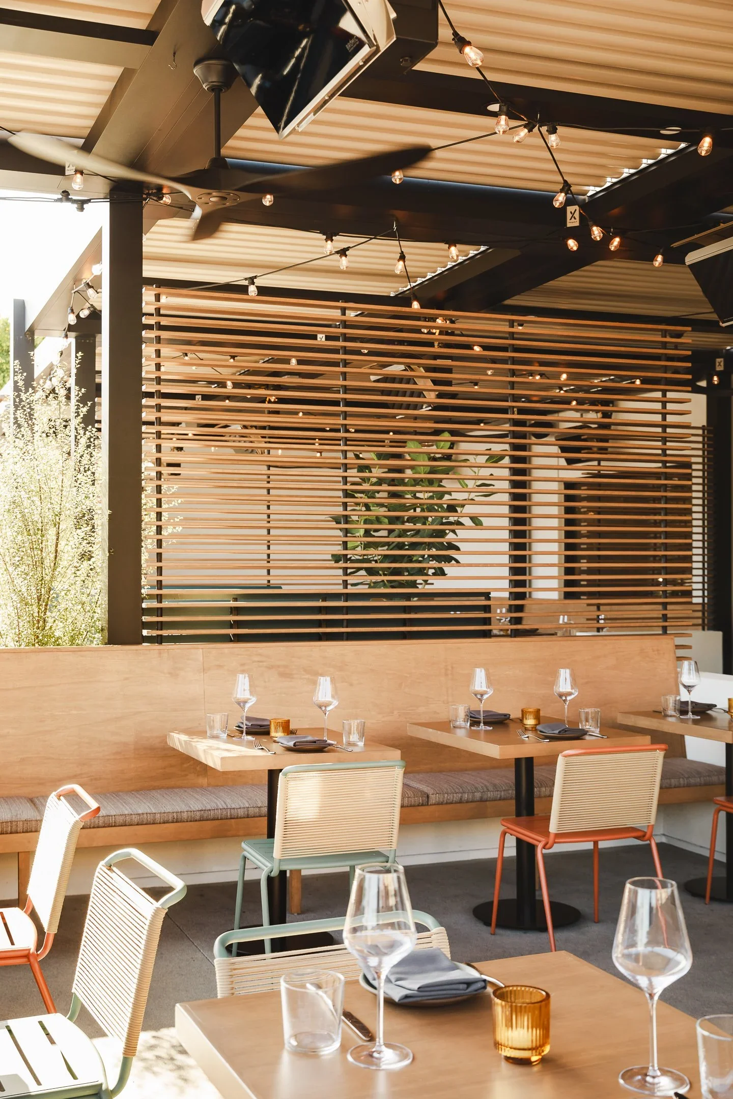 Covered outdoor patio with wood louvered privacy screen, string lights, mixed terracotta and sage rope chairs, and wood banquette seating — Marlena restaurant patio design Long Beach by Mokume Design Studio