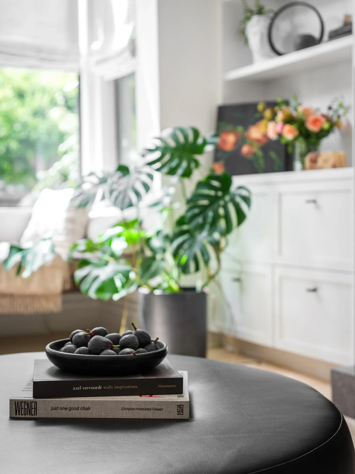 Close-up of black leather round ottoman styled with design books and ceramic bowl, with monstera plant and white built-in cabinetry in background — Pacific Heights San Francisco by Mokume Design Studio