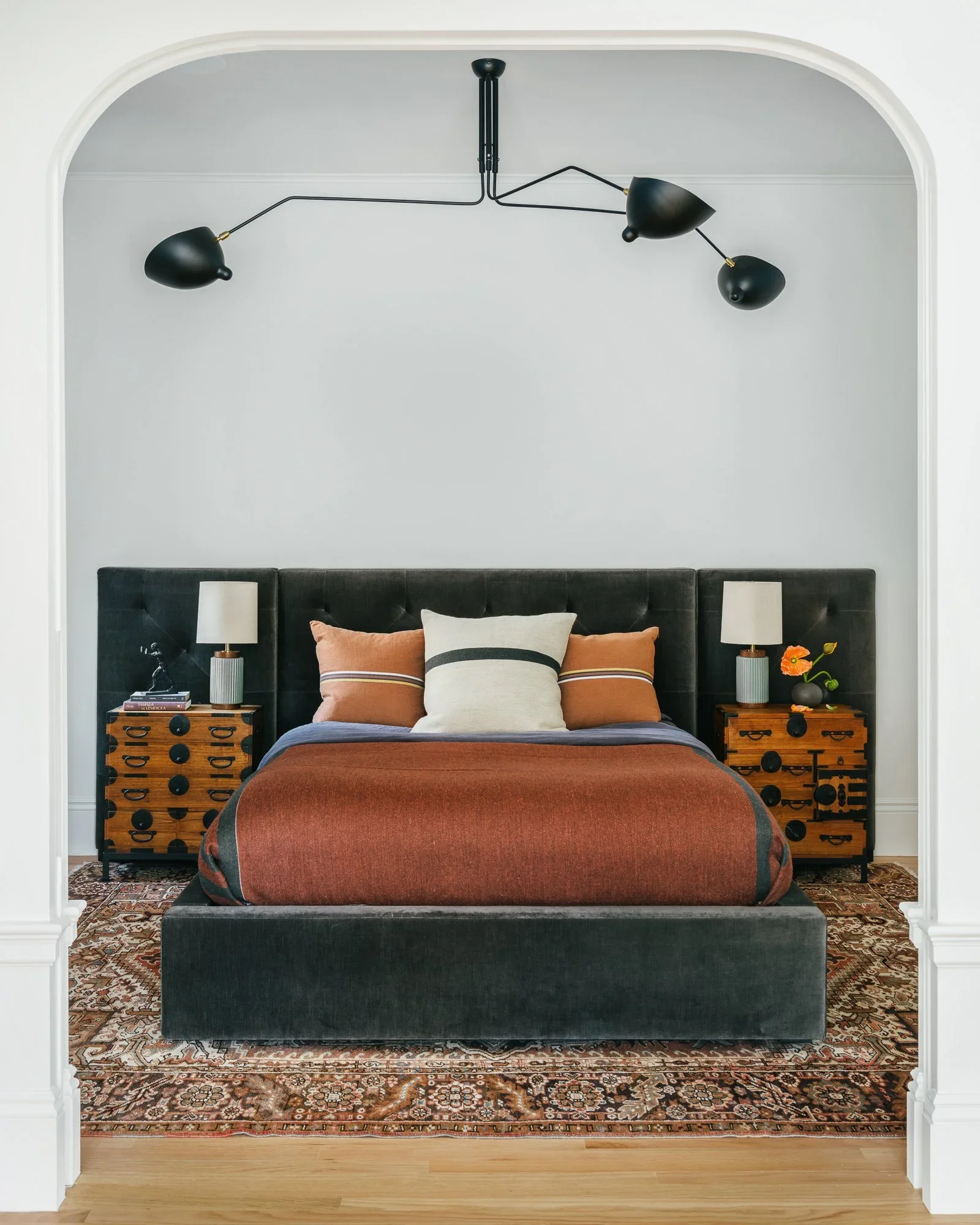 Primary bedroom framed by white plaster arch with charcoal velvet tufted headboard, Japanese tansu nightstands, Serge Mouille-style chandelier, and Persian rug — Hill Street Residence San Francisco by Mokume Design Studio