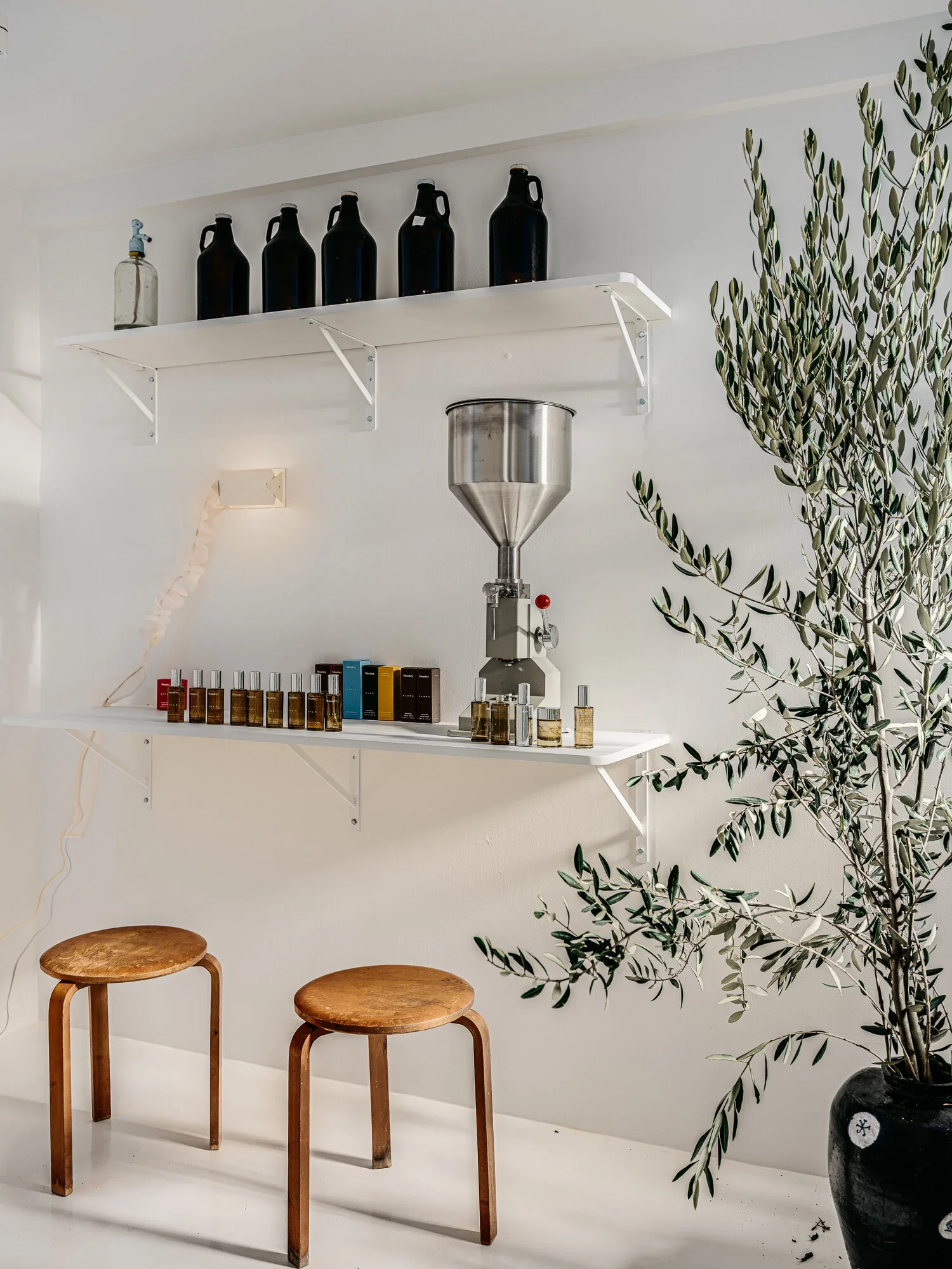 Minimalist white product dispensing station with stainless steel filler, apothecary bottles, fragrance oil display, and large potted olive tree — Monastery Made San Francisco retail design by Mokume Design Studio